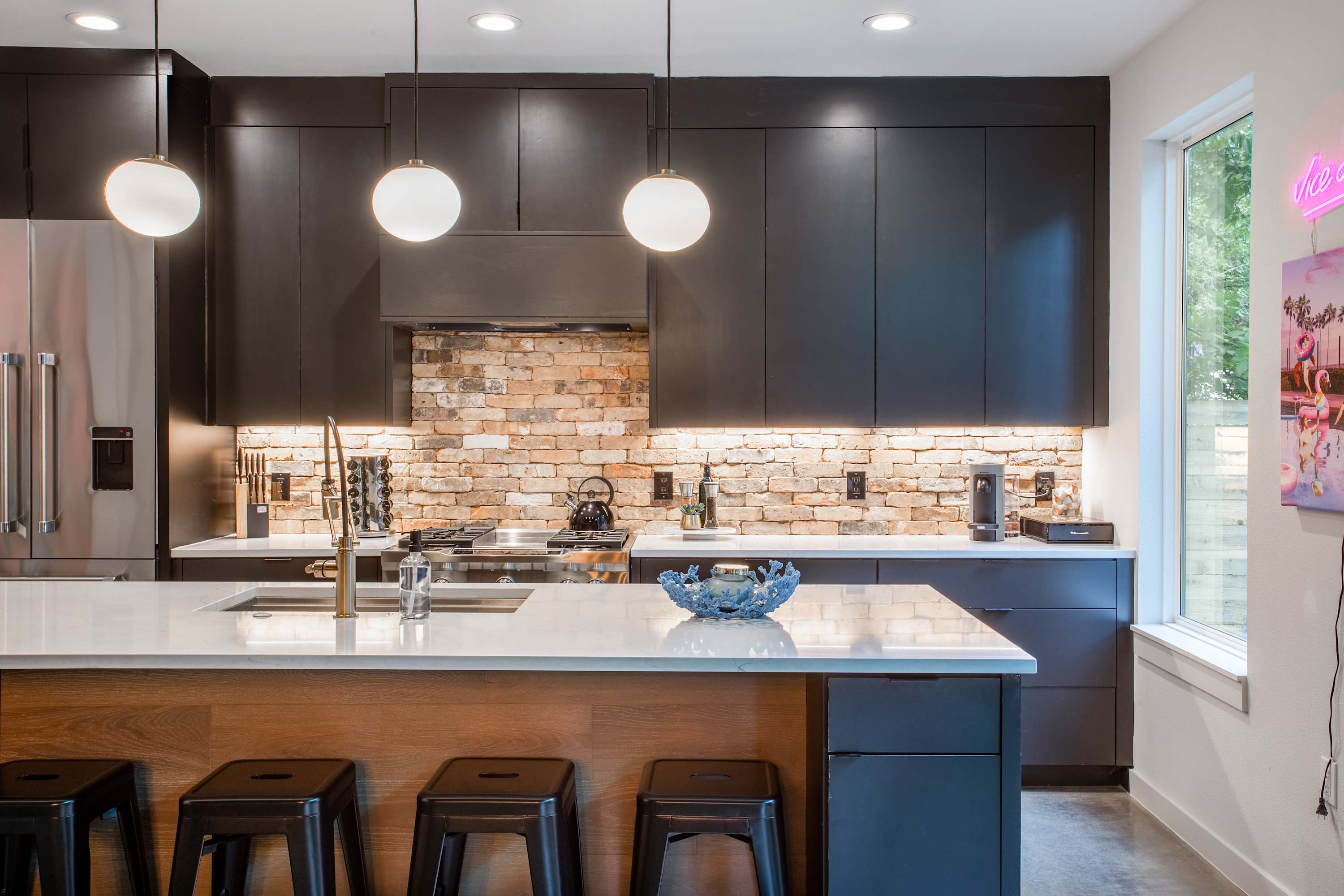 Contemporary kitchen featuring dark flat-panel cabinetry, a brick subway tile backsplash, and a large island with a white countertop and gold faucet. Under-cabinet lighting highlights the cooking area with a gas range.