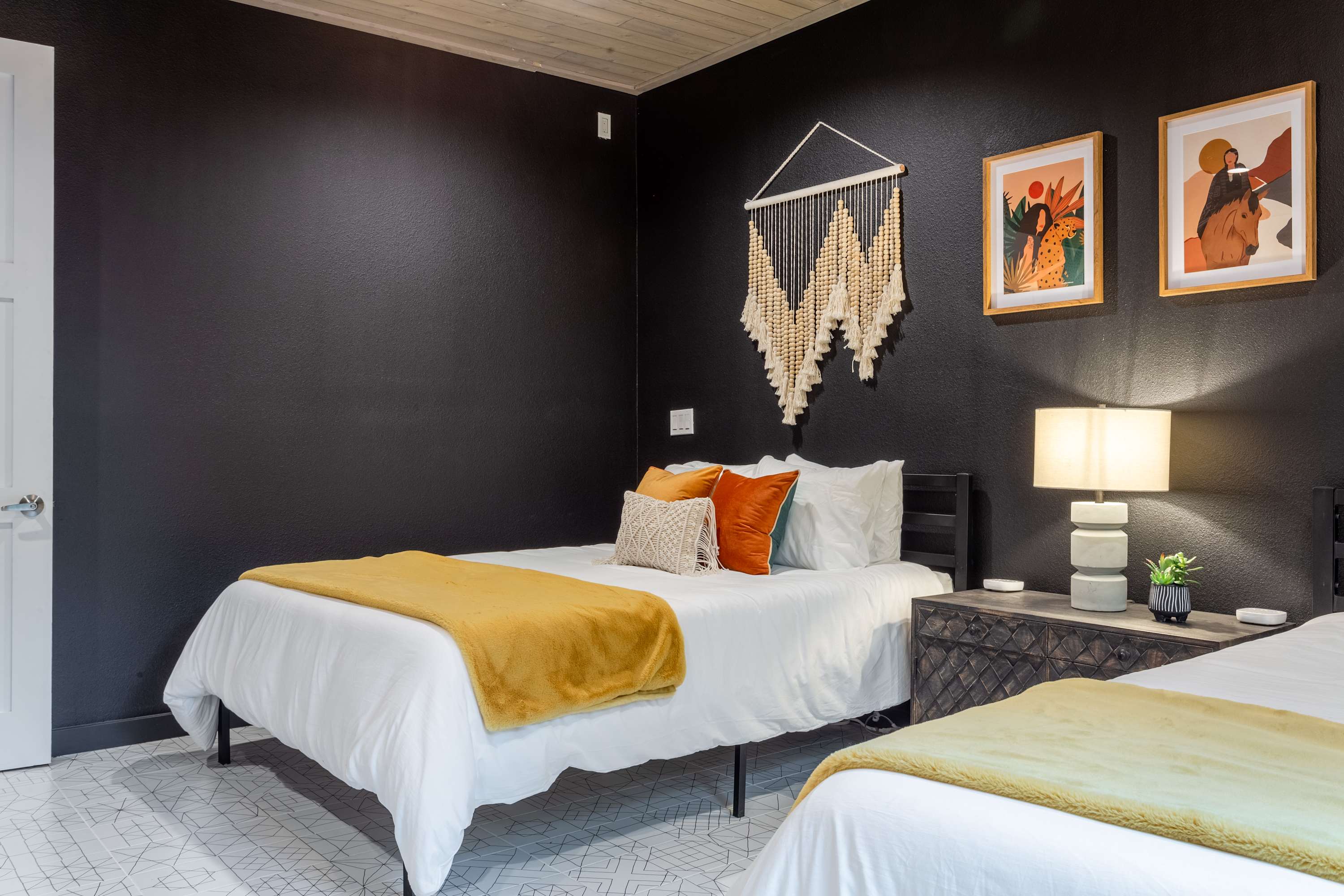 Cozy bohemian bedroom featuring a queen bed with white bedding and a mustard yellow throw, flanked by a dark wood nightstand with a lamp. Dark walls are decorated with macrame art and framed prints, creating a warm, inviting atmosphere.