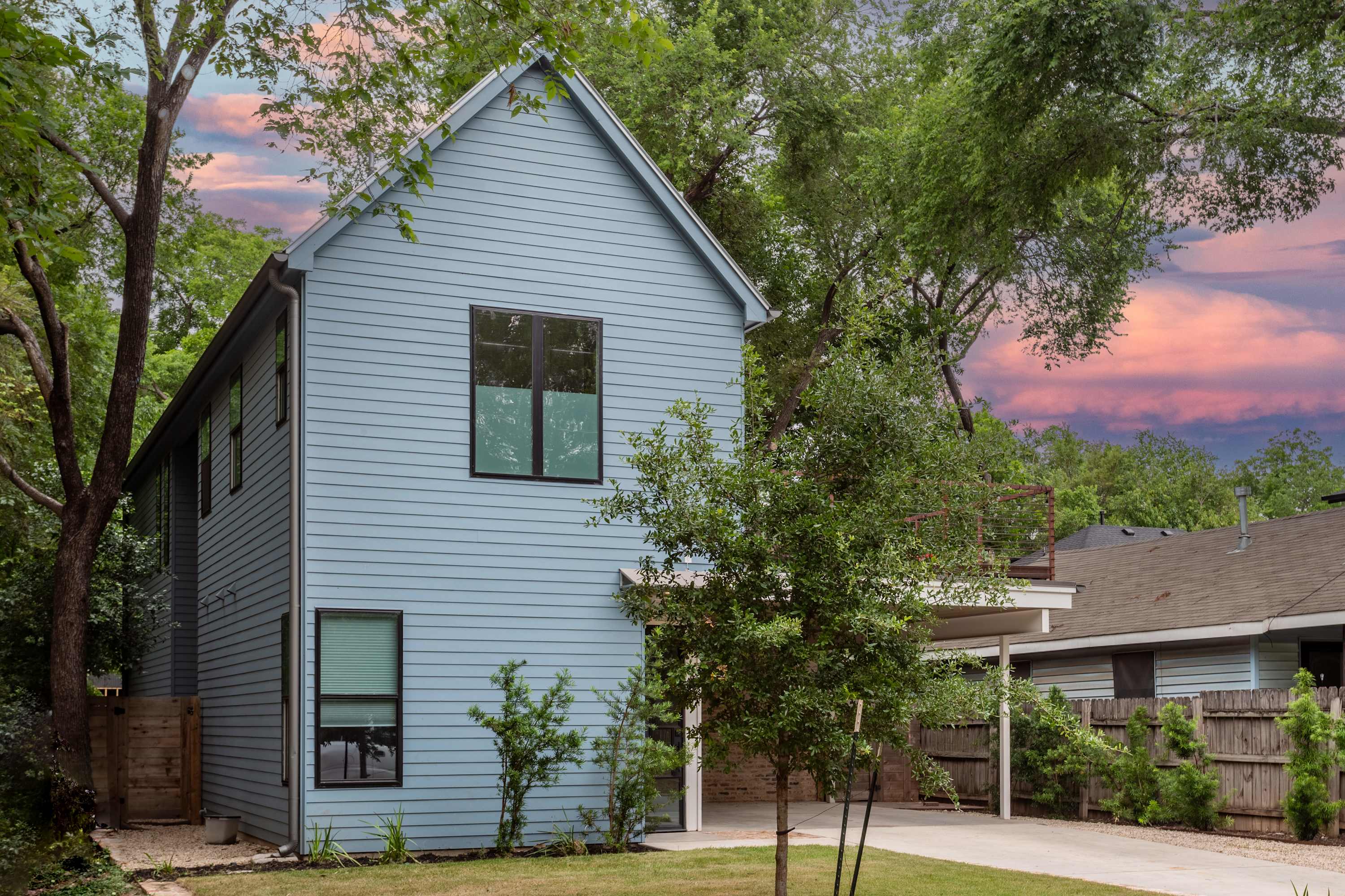 The front exterior of a modern two-story house with light blue horizontal siding and black-framed windows, surrounded by green trees and a paved driveway. The sky is a dramatic mix of blue and pink.