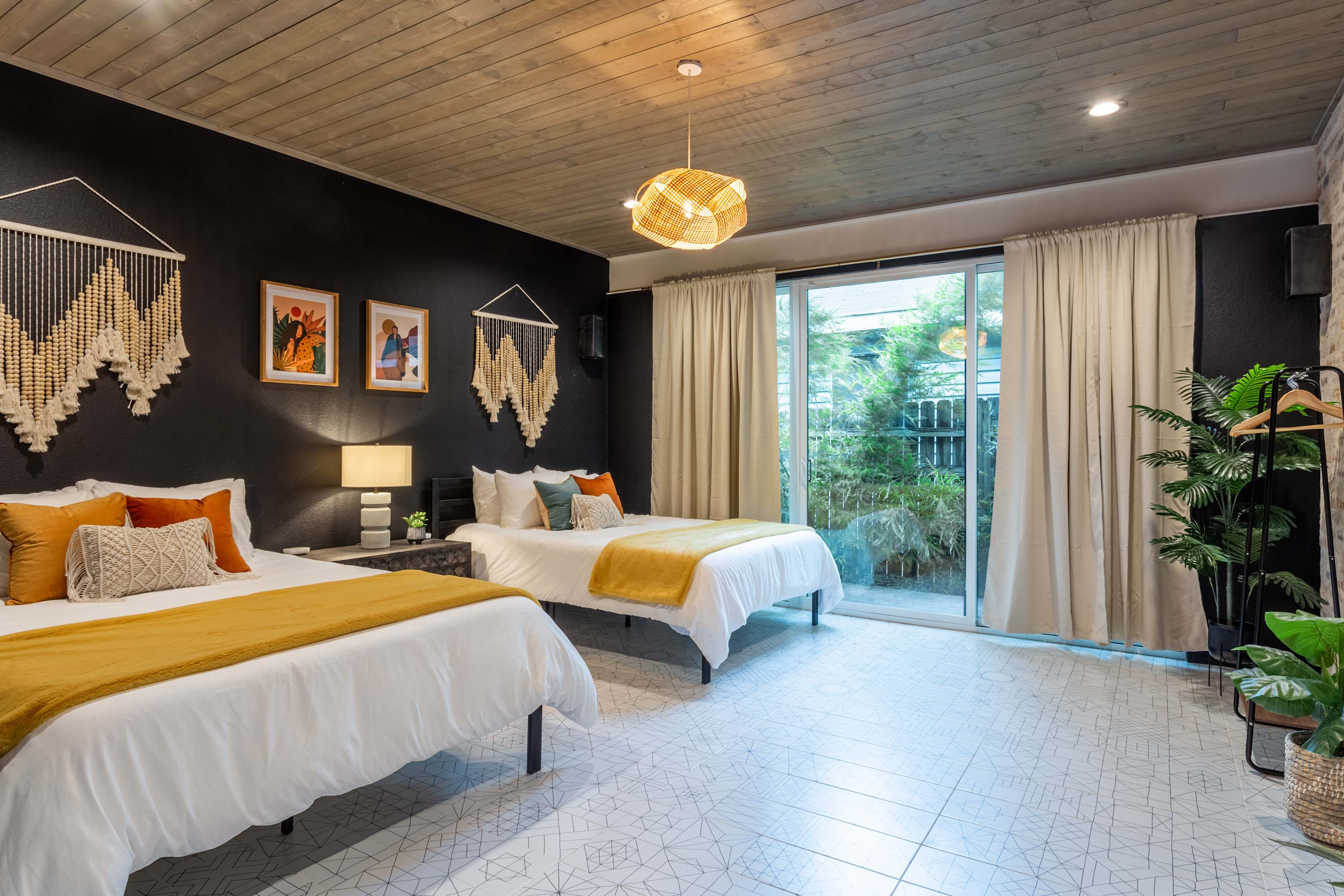 Bohemian-style bedroom with two queen beds, white bedding, and mustard yellow throws, set against dark walls. Macrame wall hangings and framed art adorn the walls, and a large sliding glass door opens to an outdoor area.