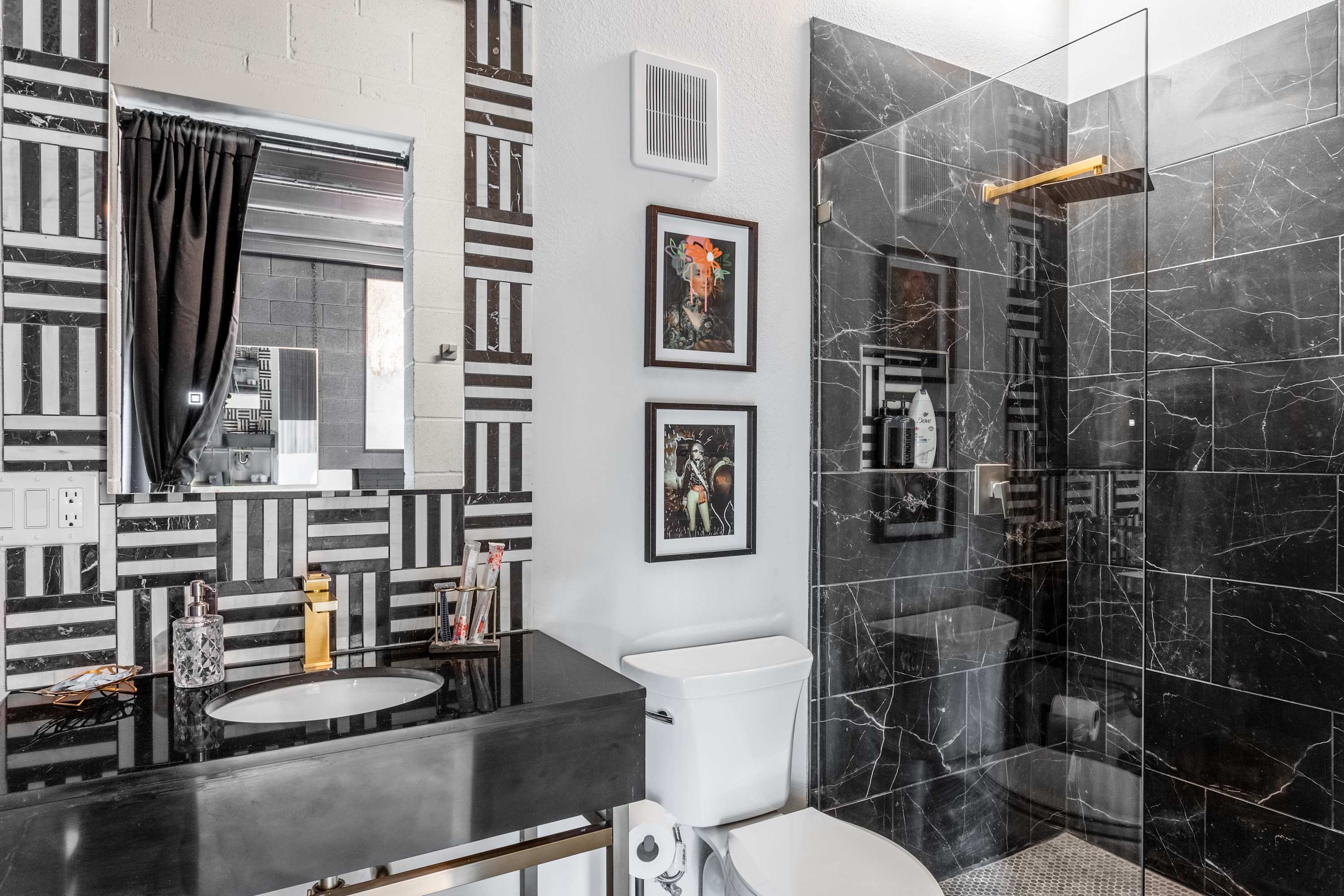 A chic bathroom showcasing a black and white geometric tiled vanity wall with a sleek black countertop and a gold faucet. The spacious walk-in shower is clad in dark marble-look tiles, complemented by a clear glass enclosure.