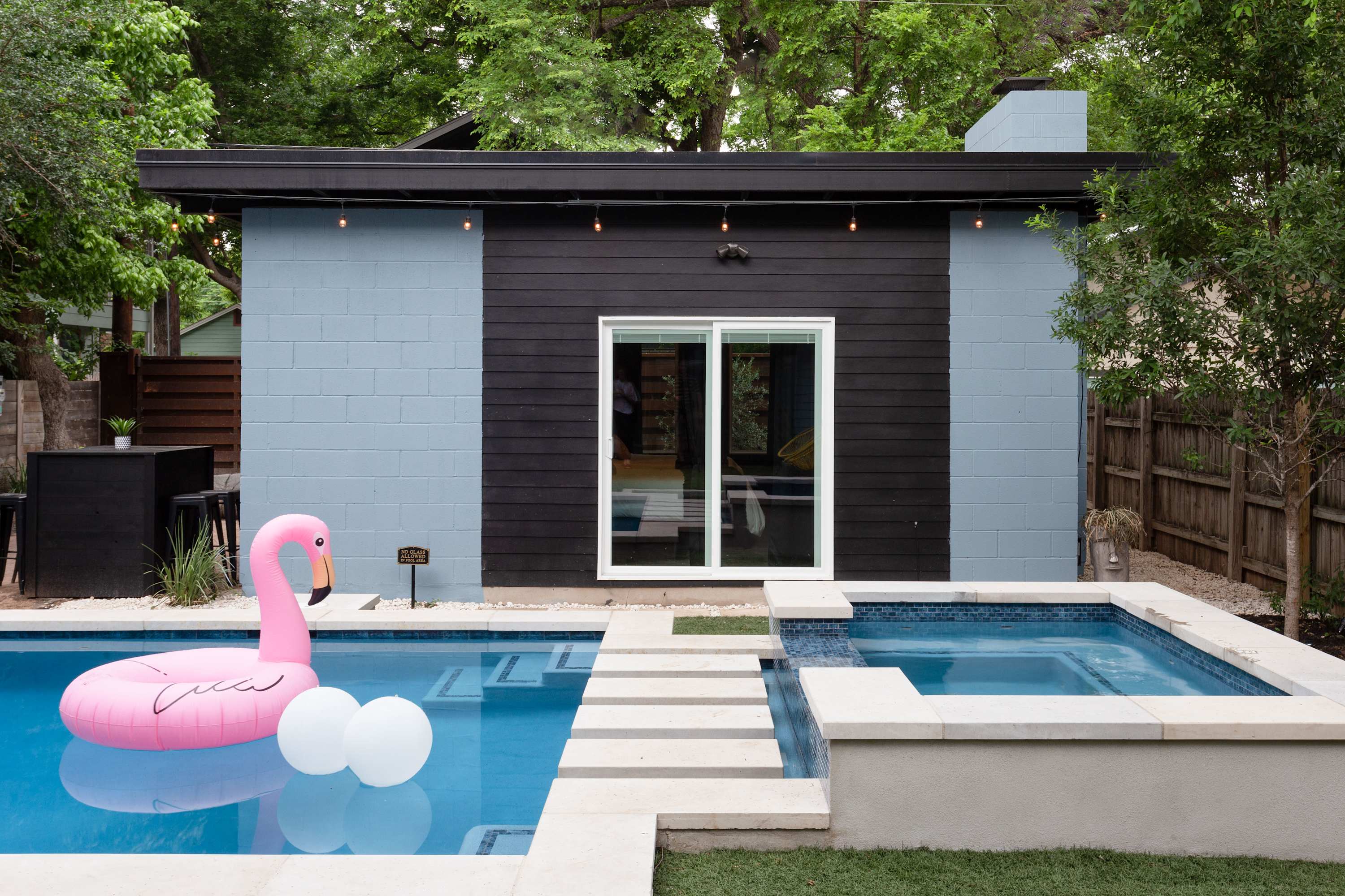 The back of a modern house with a pool and hot tub. The building features black and light blue block siding, a large sliding glass door, and a path of stepping stones leading to the pool. A pink flamingo float is in the pool.