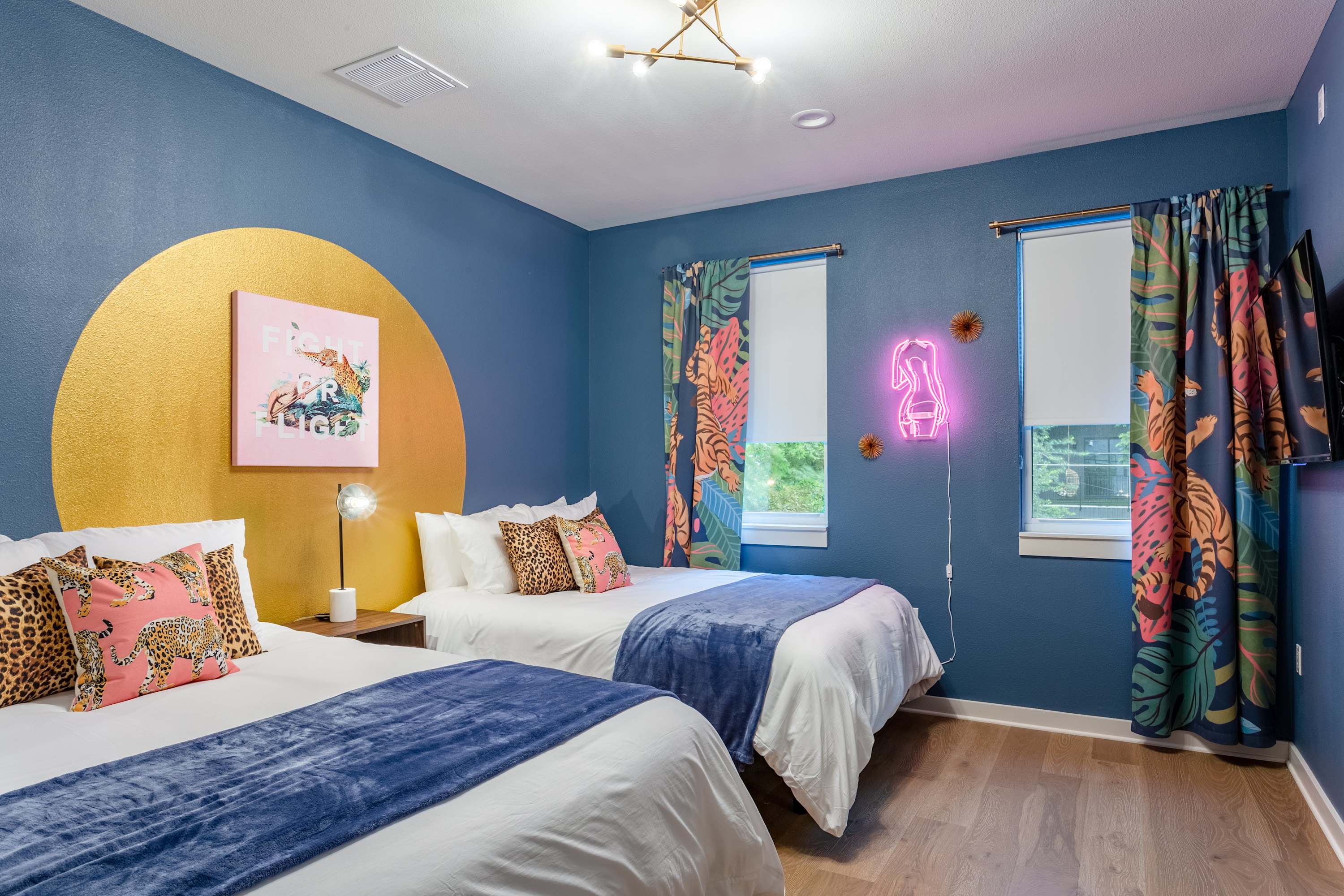 Vibrant bedroom with two queen beds, each with white bedding and a blue throw, set against a bold blue wall with a large gold circle accent. Tropical print curtains frame the windows, and a pink neon high-heel sign adds a playful touch.