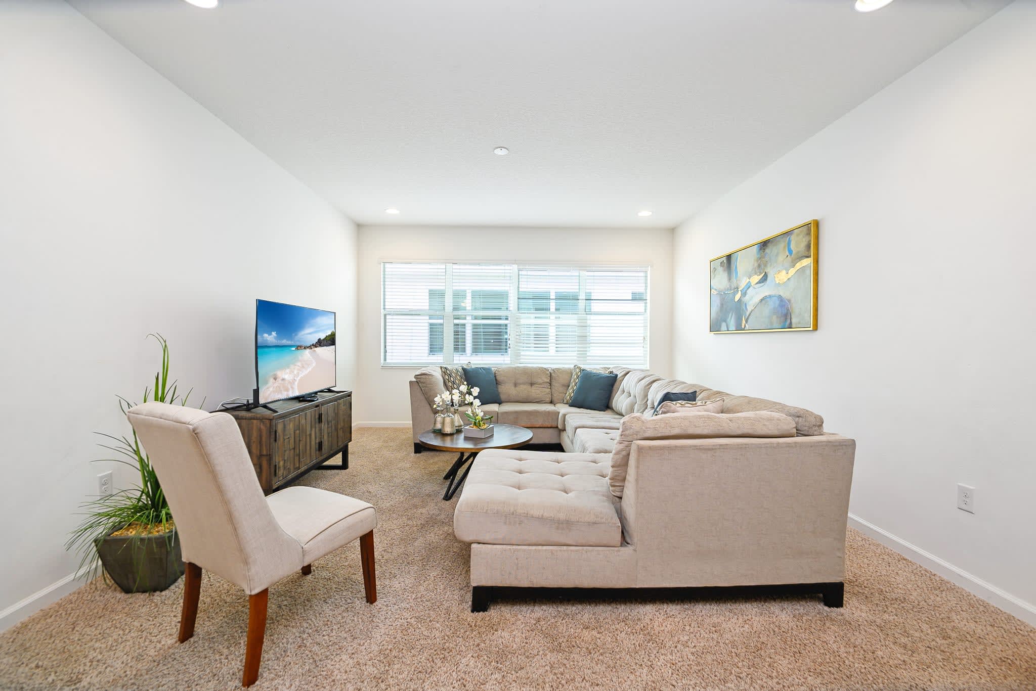 Bright living room with a large L-shaped light beige sectional sofa, a round wooden coffee table, and a TV on a rustic wooden console. Large windows with blinds provide natural light, enhancing the airy feel of the room.