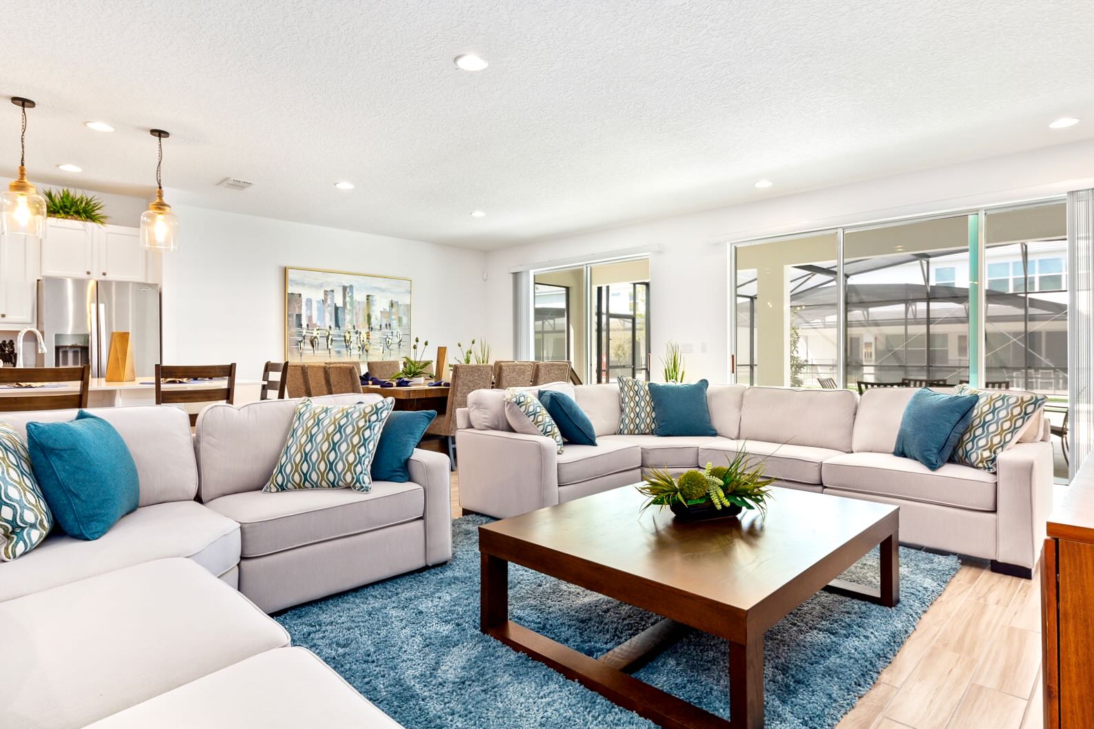 Bright living room featuring a large light-colored sectional sofa with blue and patterned throw pillows, a dark wood coffee table, and a blue shag rug, with sliding glass doors opening to a screened patio and pool.