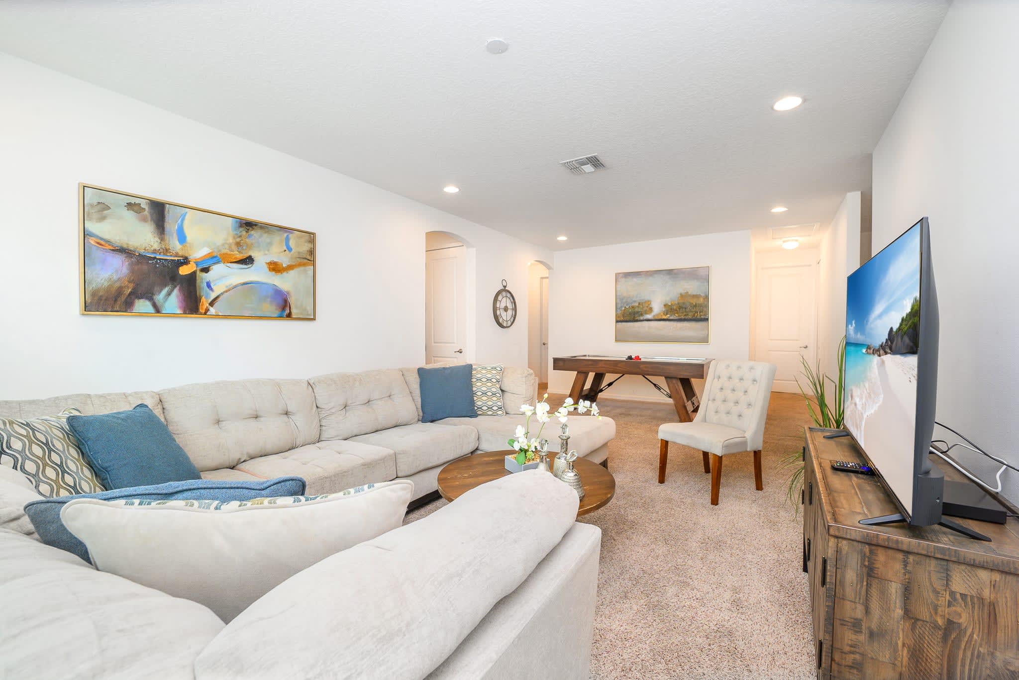 Cozy living area featuring a large L-shaped light beige sectional sofa with blue throw pillows, a round wooden coffee table, and a wall-mounted TV. A pool table is visible in the background, suggesting a multi-functional space.