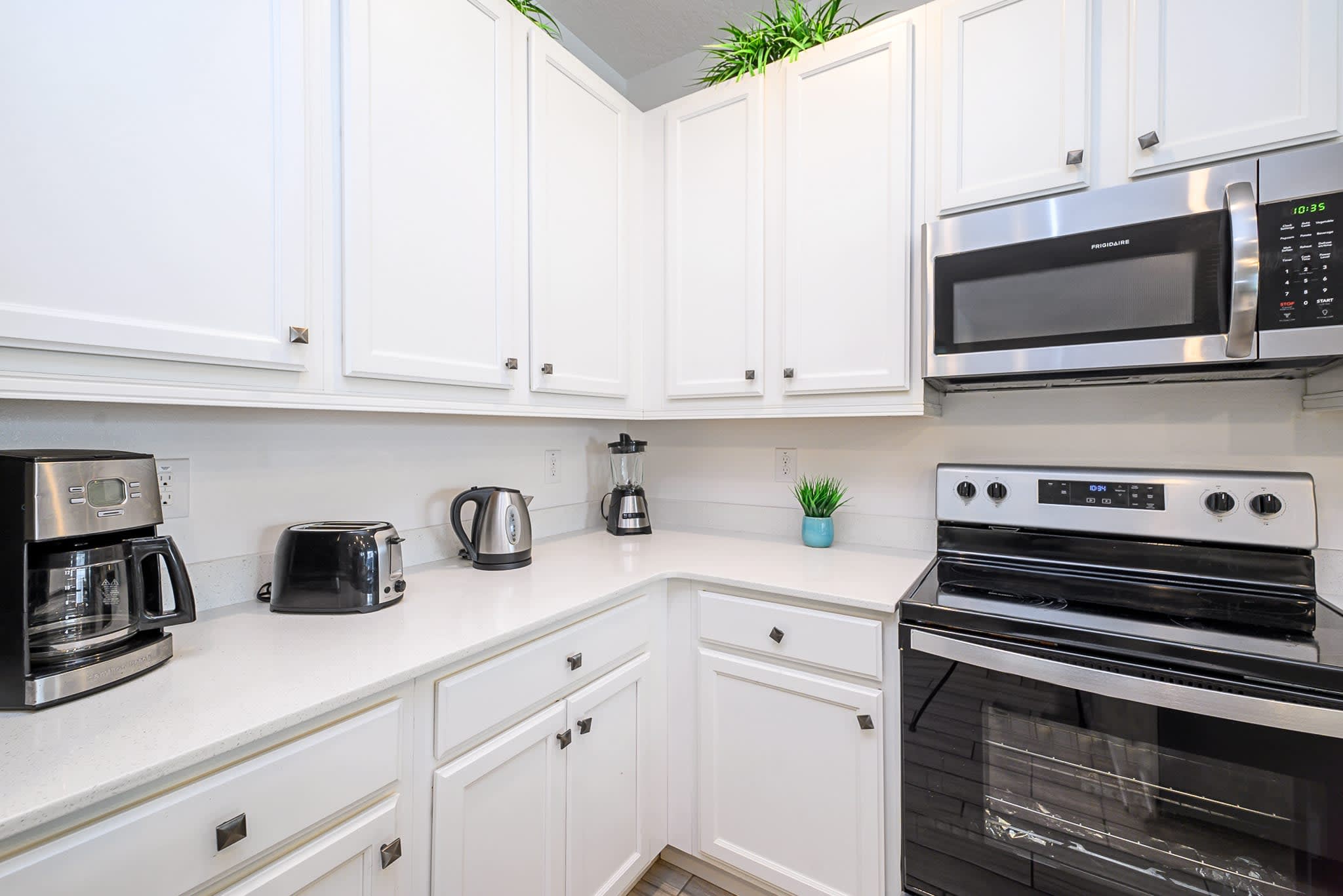 Corner of a modern kitchen counter with white quartz countertops and white cabinets, equipped with a coffee maker, toaster, blender, and a black electric range with an overhead microwave.