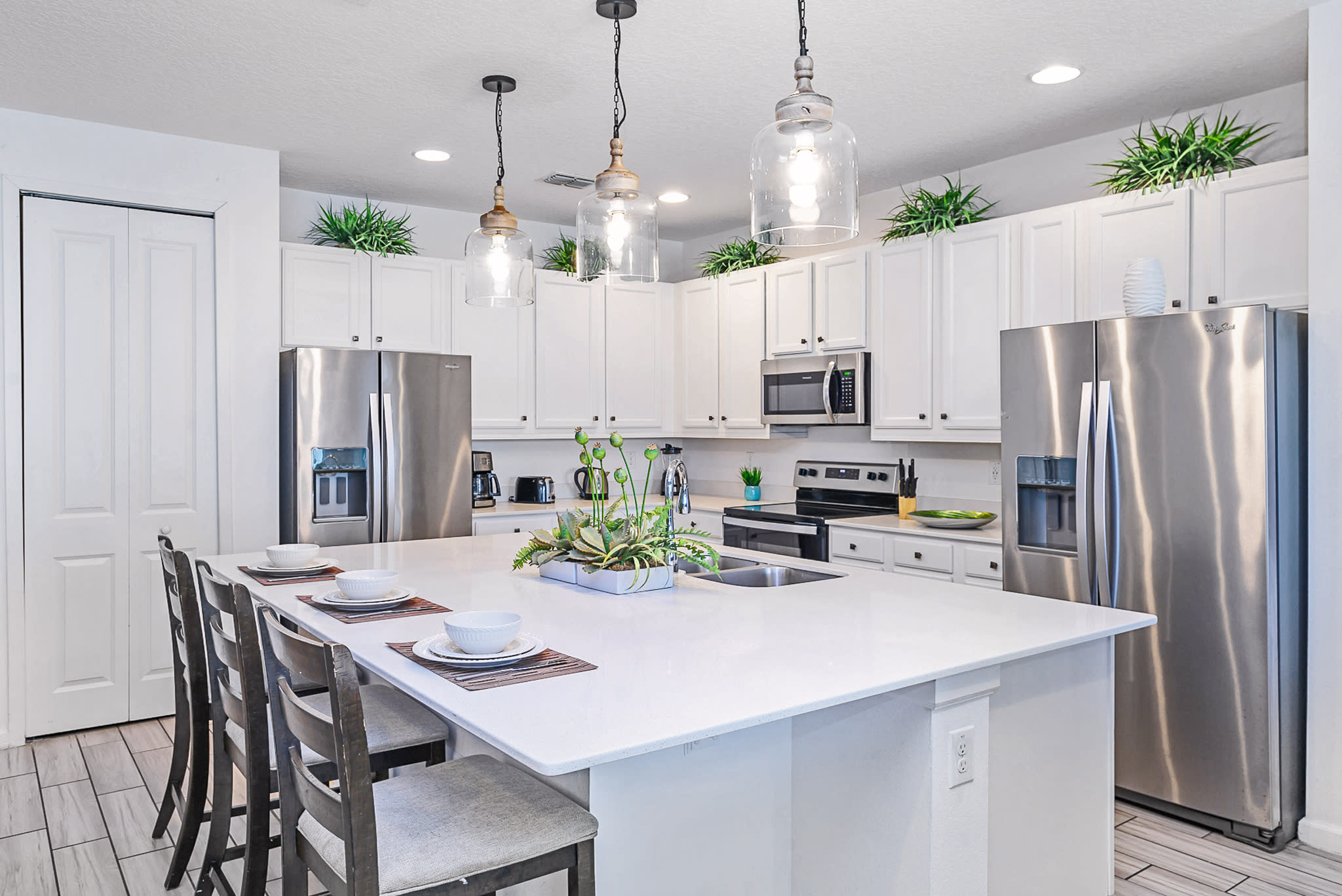 Modern kitchen featuring white shaker cabinets, stainless steel appliances including two refrigerators, and a large white quartz island with seating for four, illuminated by pendant lights.