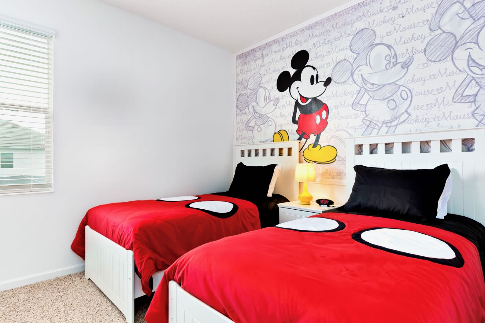 A vibrant children's bedroom with a Mickey Mouse theme, featuring two twin beds with red and black bedding. A large Mickey Mouse mural adorns the wall above the white headboards.