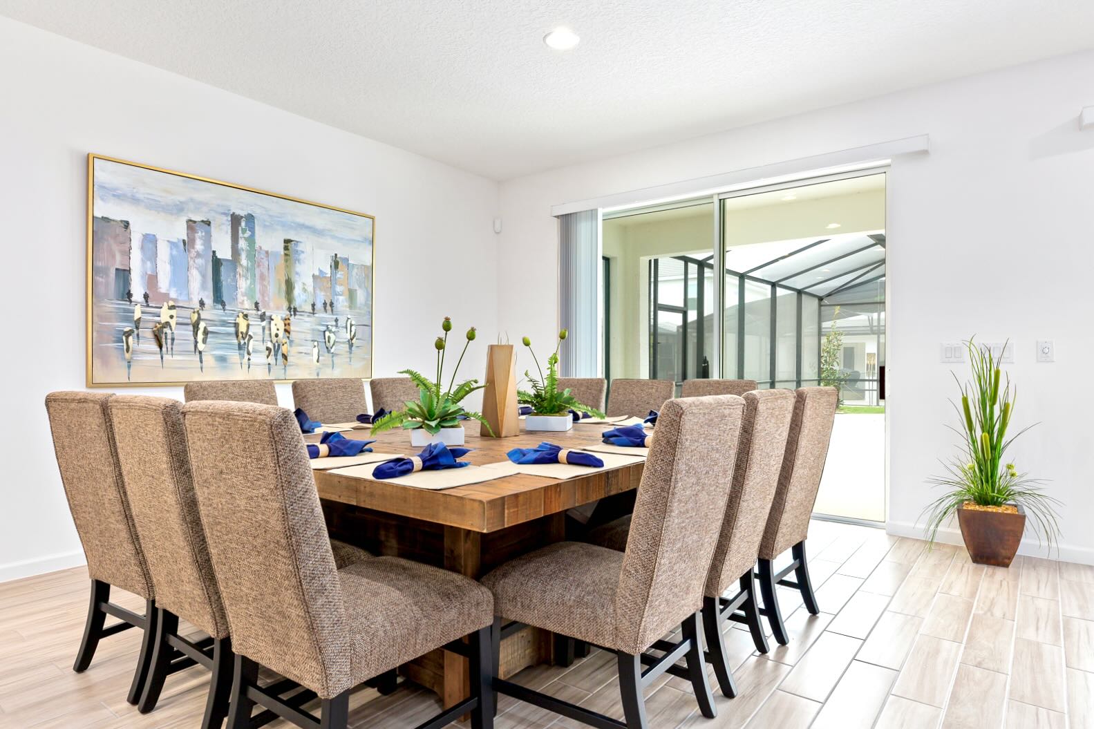 Spacious dining room with a large square rustic wooden table surrounded by eight upholstered chairs, set with blue napkins, and featuring a modern cityscape painting on the wall.