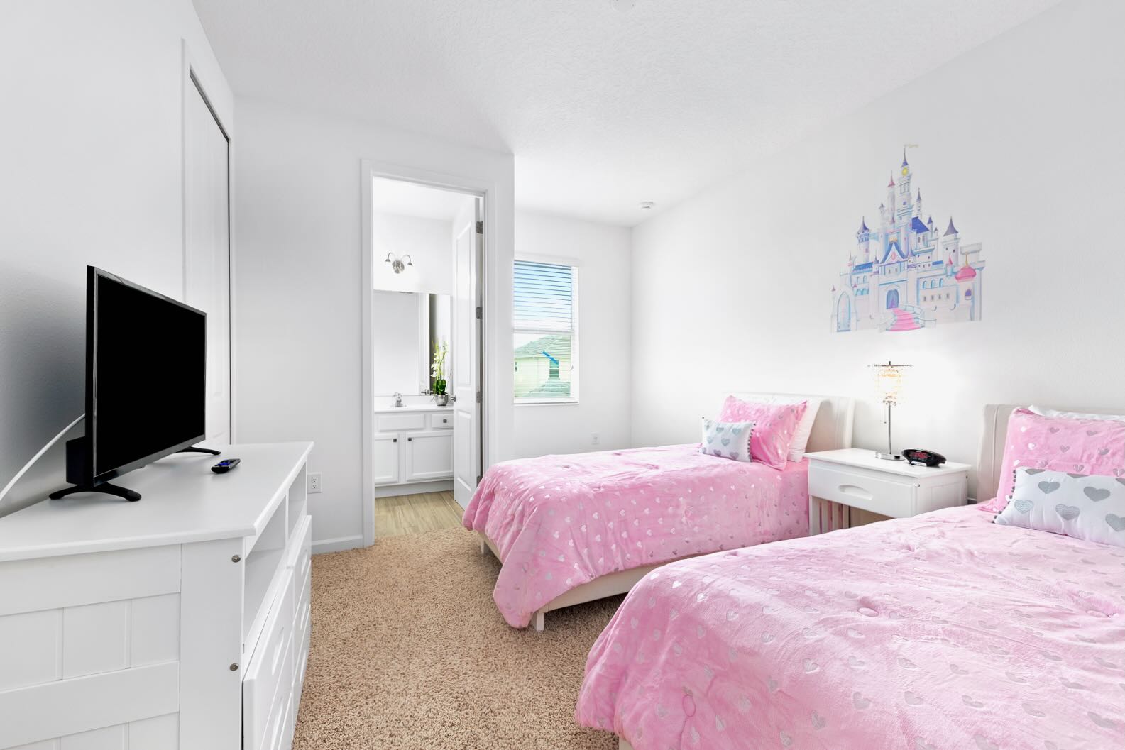 A bright children's bedroom featuring two twin beds with pink heart-patterned bedding, a white dresser with a TV, and a large castle wall decal. An open doorway leads to an en-suite bathroom.
