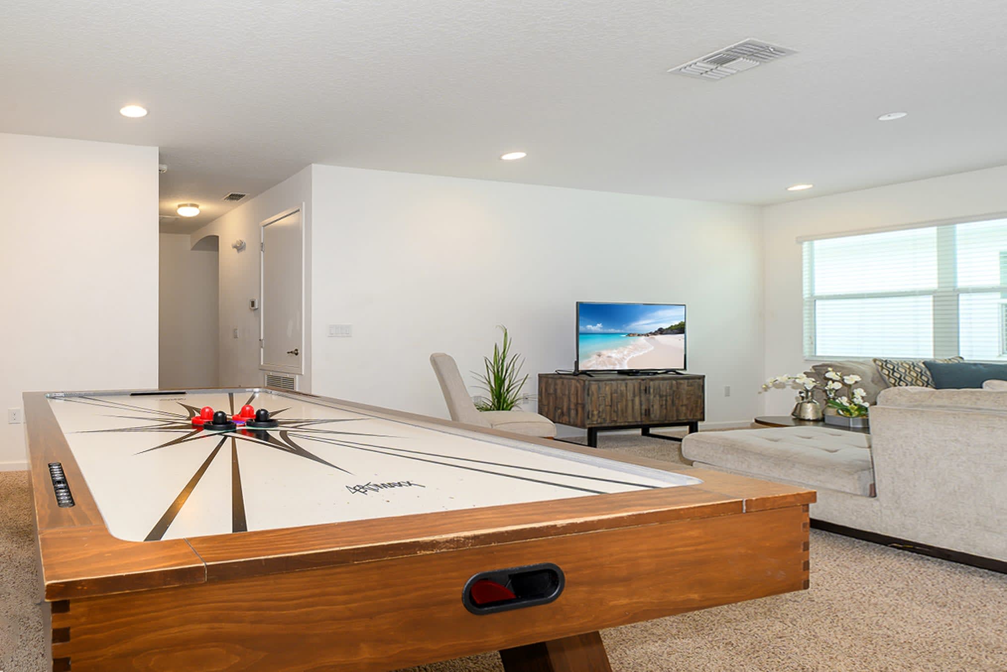 Bright game room featuring a wooden air hockey table with a black and white star design, a wall-mounted TV on a rustic wooden console, and a light-colored sectional sofa in the background. The room has a cozy, carpeted floor.