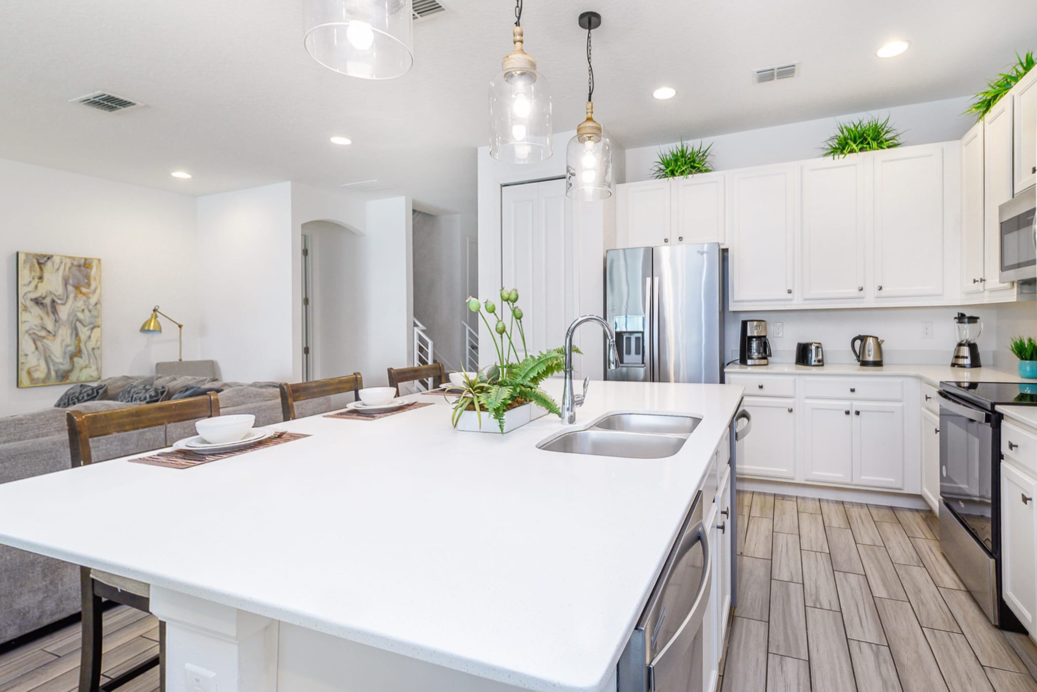 Modern open-concept kitchen featuring white shaker cabinets, stainless steel appliances, a large white quartz island with a double sink, and three pendant lights. The adjacent living area is visible in the background.