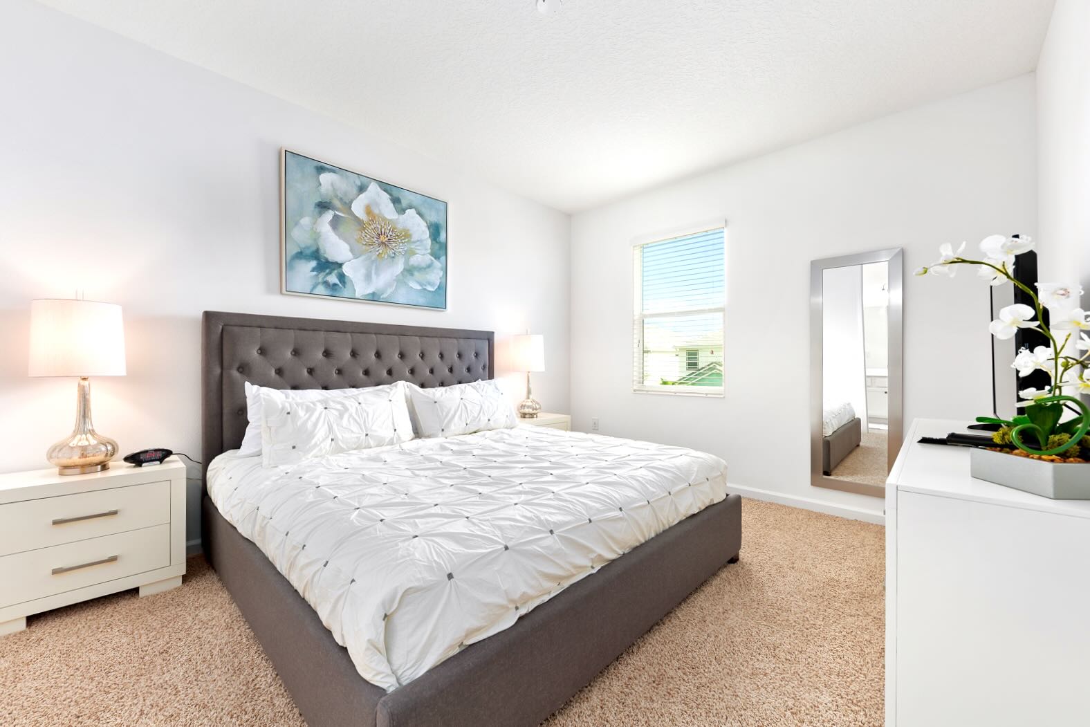 Bright bedroom with a large gray upholstered king-size bed, crisp white bedding, and two white nightstands with lamps. A large floral artwork hangs above the bed, complemented by a full-length mirror and a window with blinds.