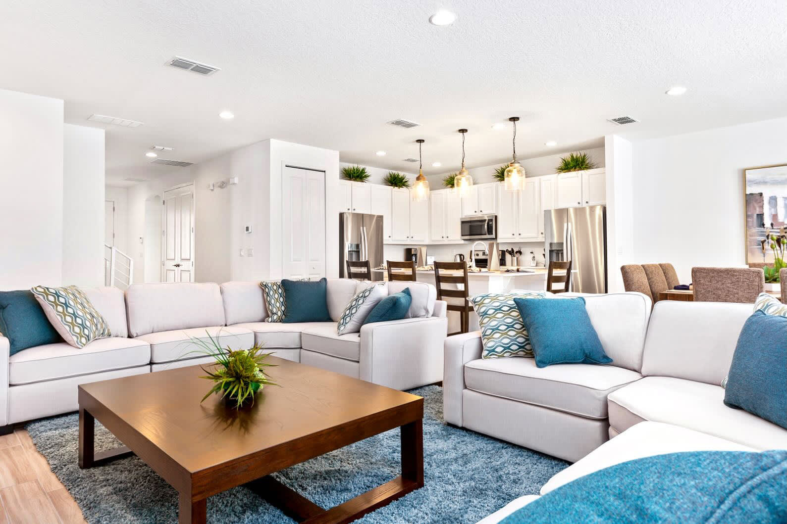 Spacious open-concept living room with a large L-shaped white sectional sofa, teal and patterned throw pillows, a dark wood coffee table, and a blue area rug. The kitchen is visible in the background, creating a cohesive living space.