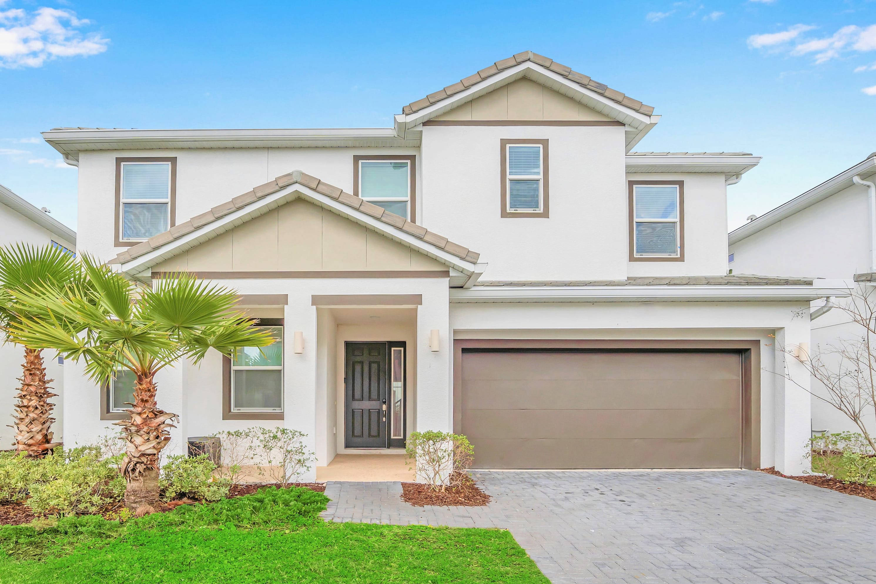 A modern two-story house with a white stucco exterior, brown trim, and a dark brown garage door. Palm trees and green landscaping adorn the front, with a paved driveway leading to the entrance.