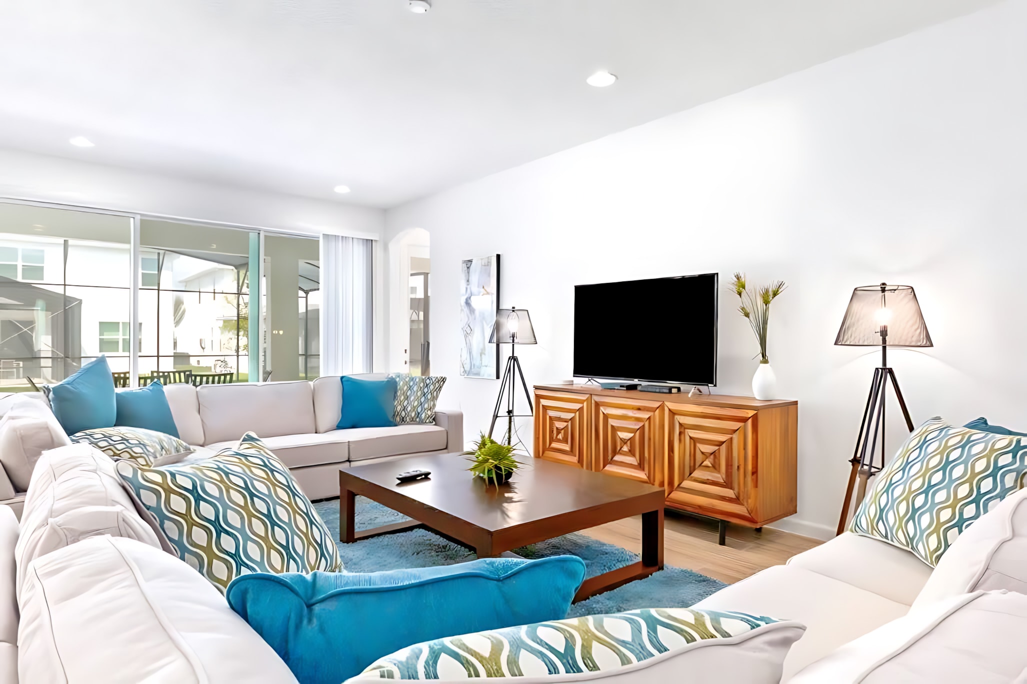 Bright living room with a large light-colored sectional sofa adorned with blue and patterned pillows, a dark wood coffee table, a TV on a wooden console, and sliding glass doors leading to the screened pool area.