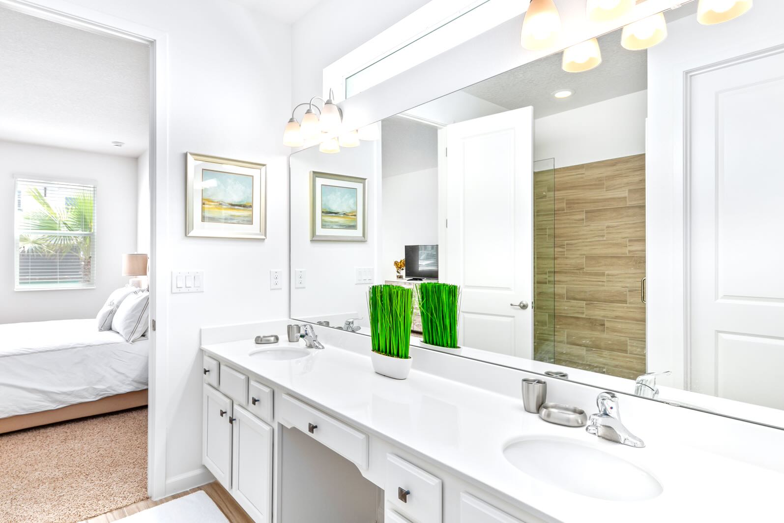 Bright bathroom featuring a long white vanity with double sinks, a large mirror, and modern light fixtures. The walk-in shower with wood-look tile is visible through the doorway, connecting to a bedroom.