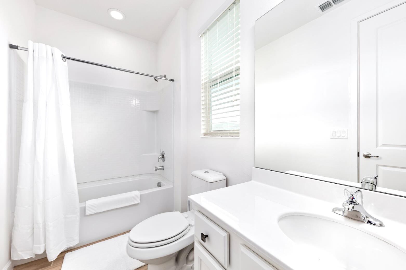 A bright, minimalist bathroom with a white bathtub and shower, a toilet, and a single vanity with a large mirror. A window with blinds provides natural light, and the floor is light wood-look tile.