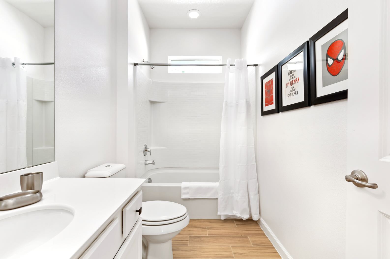 A clean, modern bathroom featuring a bathtub with a shower, a toilet, and a single vanity with a large mirror. The wall is decorated with three framed Spider-Man themed art pieces.