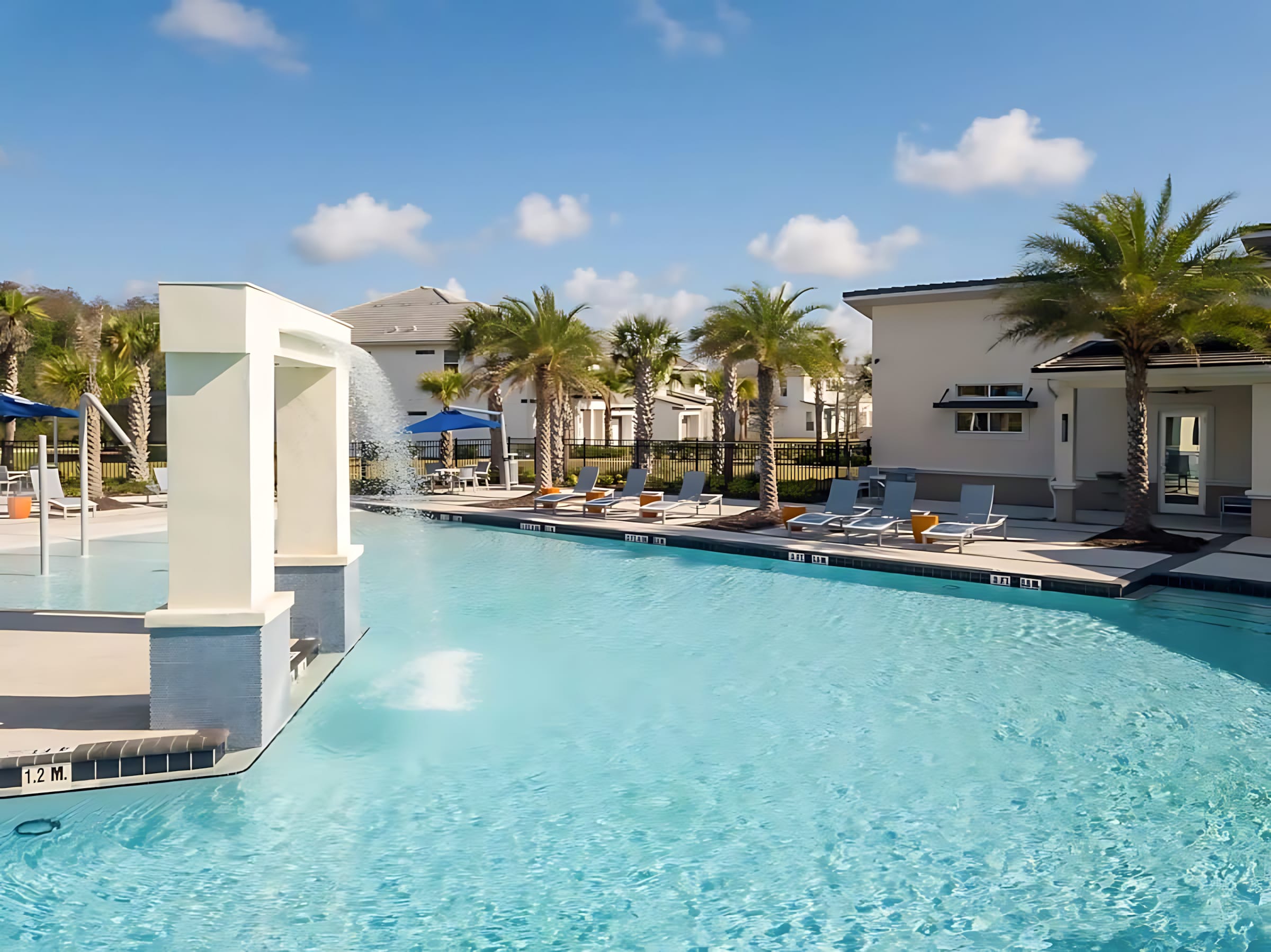 A large, bright blue communal swimming pool with a modern white water feature, surrounded by lounge chairs and palm trees under a clear sky. Residential buildings are visible in the background.