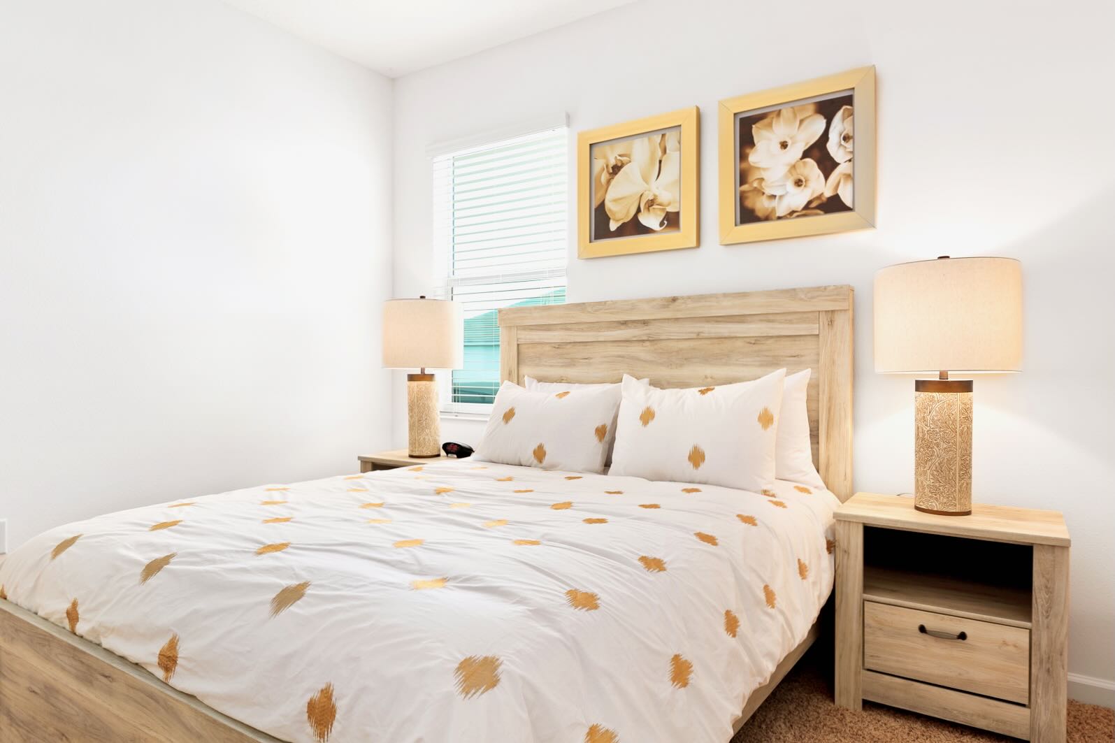 Cozy bedroom featuring a light wood queen-size bed with white and gold patterned bedding, flanked by matching nightstands and lamps. Two framed floral artworks adorn the wall above the headboard, and a window with blinds provides natural light.