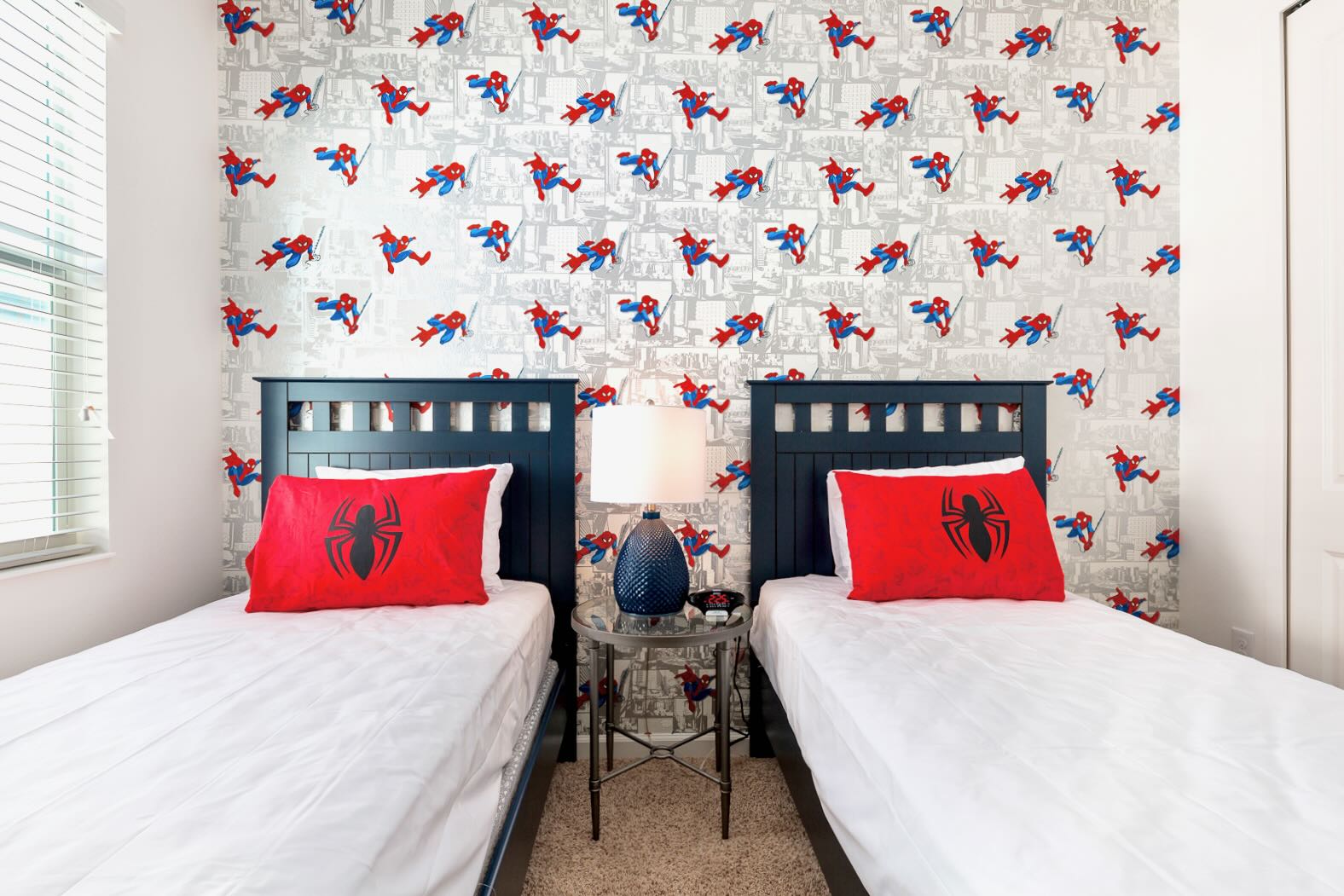 A themed children's bedroom featuring two twin beds with dark blue headboards and Spider-Man patterned wallpaper. Red pillows with a spider emblem add to the superhero decor.