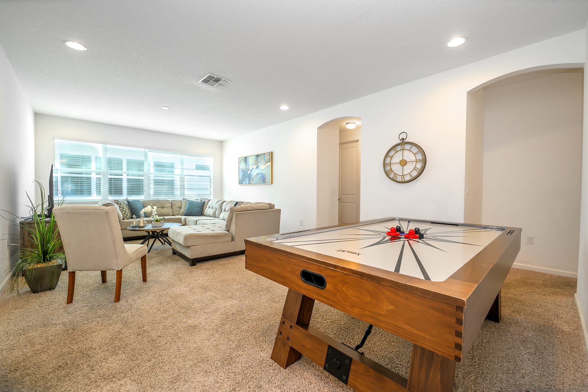 Game room featuring a wooden air hockey table in the foreground, with a cozy seating area including a sectional sofa and coffee table visible through an archway, all on light brown carpet.