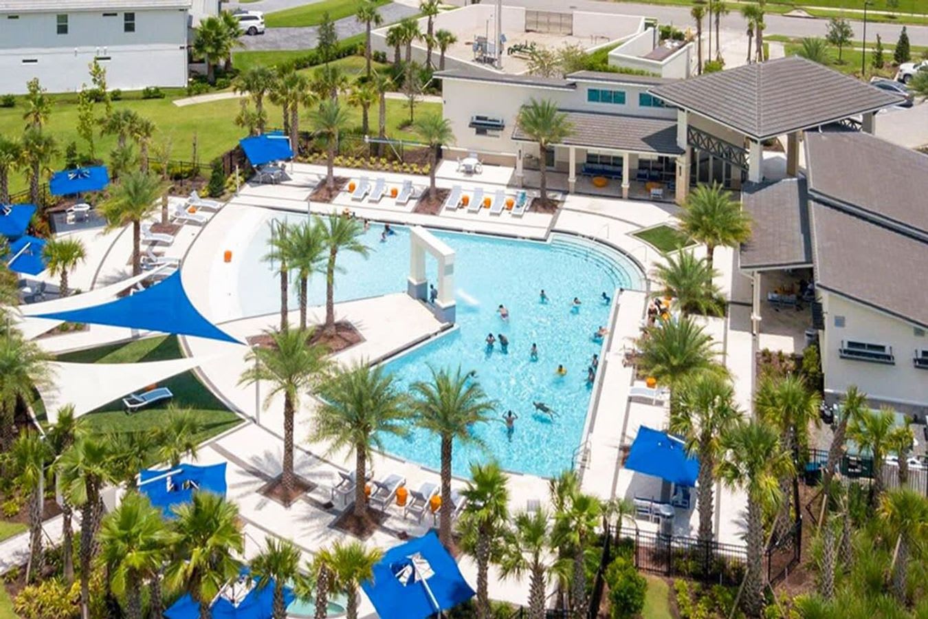 An aerial view of a large, resort-style communal swimming pool with multiple lounge chairs, blue umbrellas, and palm trees. A modern clubhouse building is visible in the background.