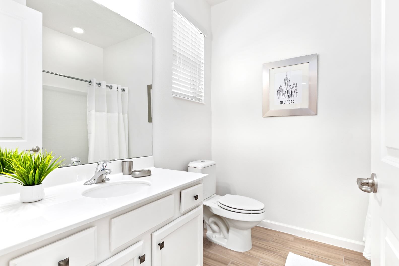 Modern white bathroom with a single vanity, a large mirror, and a bathtub with a shower curtain. A framed 'New York' artwork hangs above the toilet, and wood-look tile flooring complements the clean aesthetic.