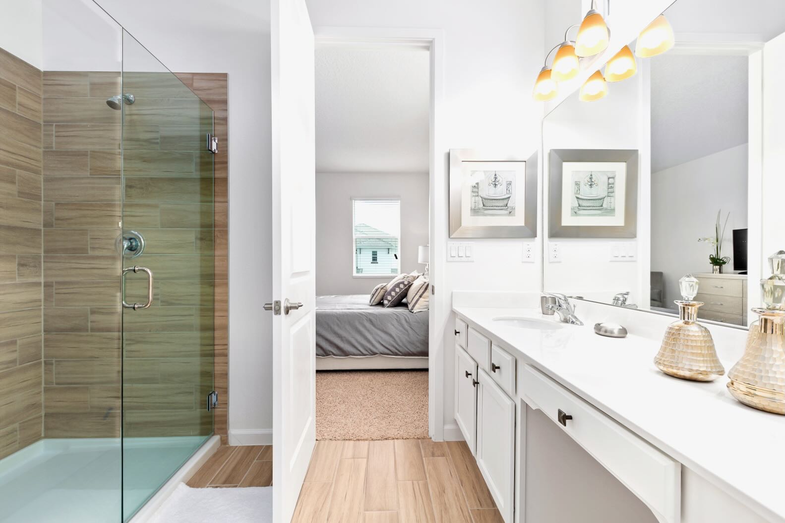 Modern bathroom featuring a glass-enclosed walk-in shower with wood-look tile walls, a white vanity with a single sink, and warm pendant lights. A bedroom is visible through the open doorway, connecting the spaces.