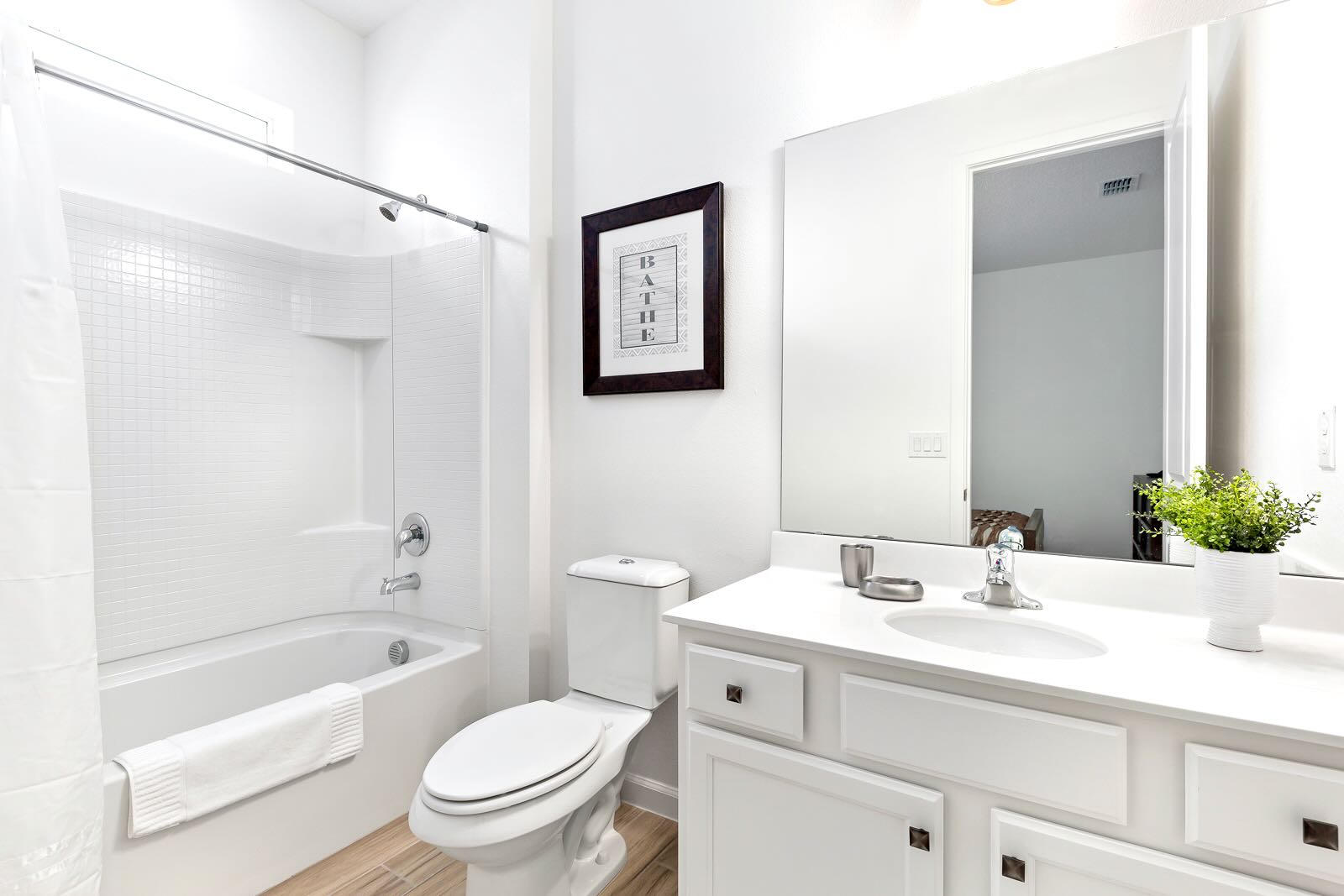 A clean, white bathroom featuring a bathtub with a shower, a toilet, and a single vanity with a large mirror. The floor is light wood-look tile, and a framed "BATHE" sign hangs on the wall.