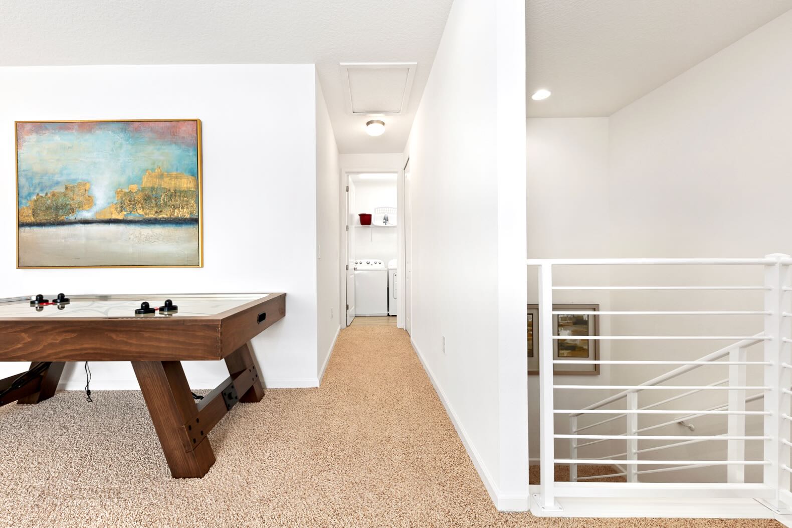 View of a game room with a wooden air hockey table, leading into a carpeted hallway with a laundry closet at the end. A modern white staircase railing is visible on the right, connecting different levels of the home.