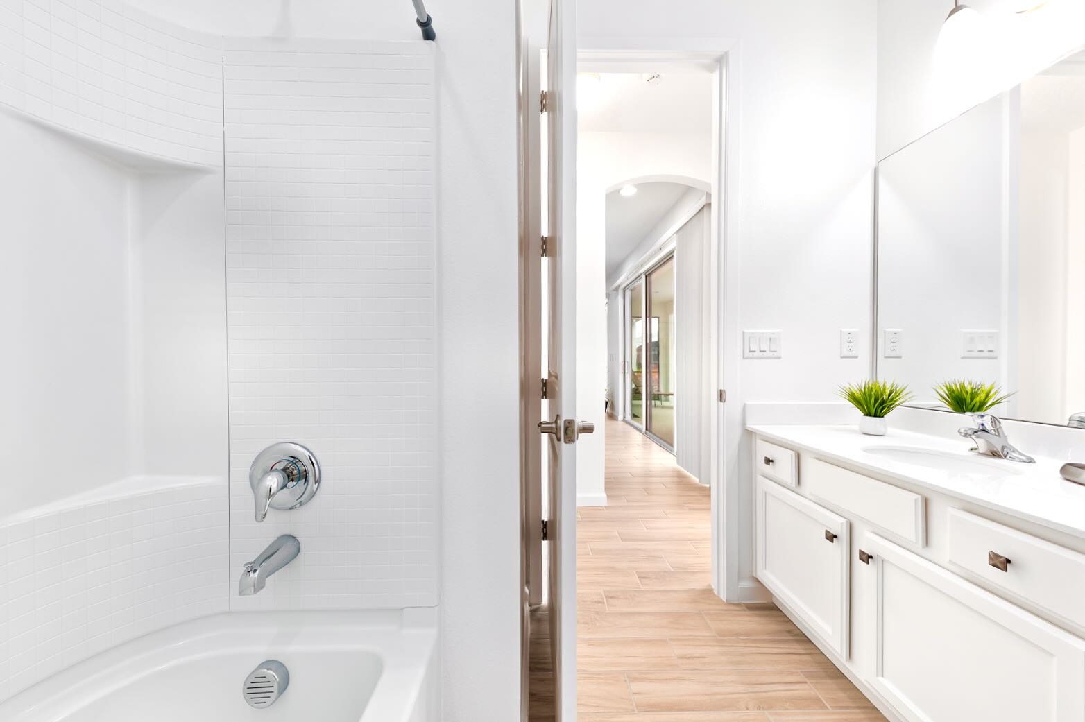 Bright bathroom featuring a white double vanity with two sinks and a large mirror, alongside a bathtub with a shower curtain. Wood-look tile flooring extends into the hallway, and a small plant adds a touch of greenery.