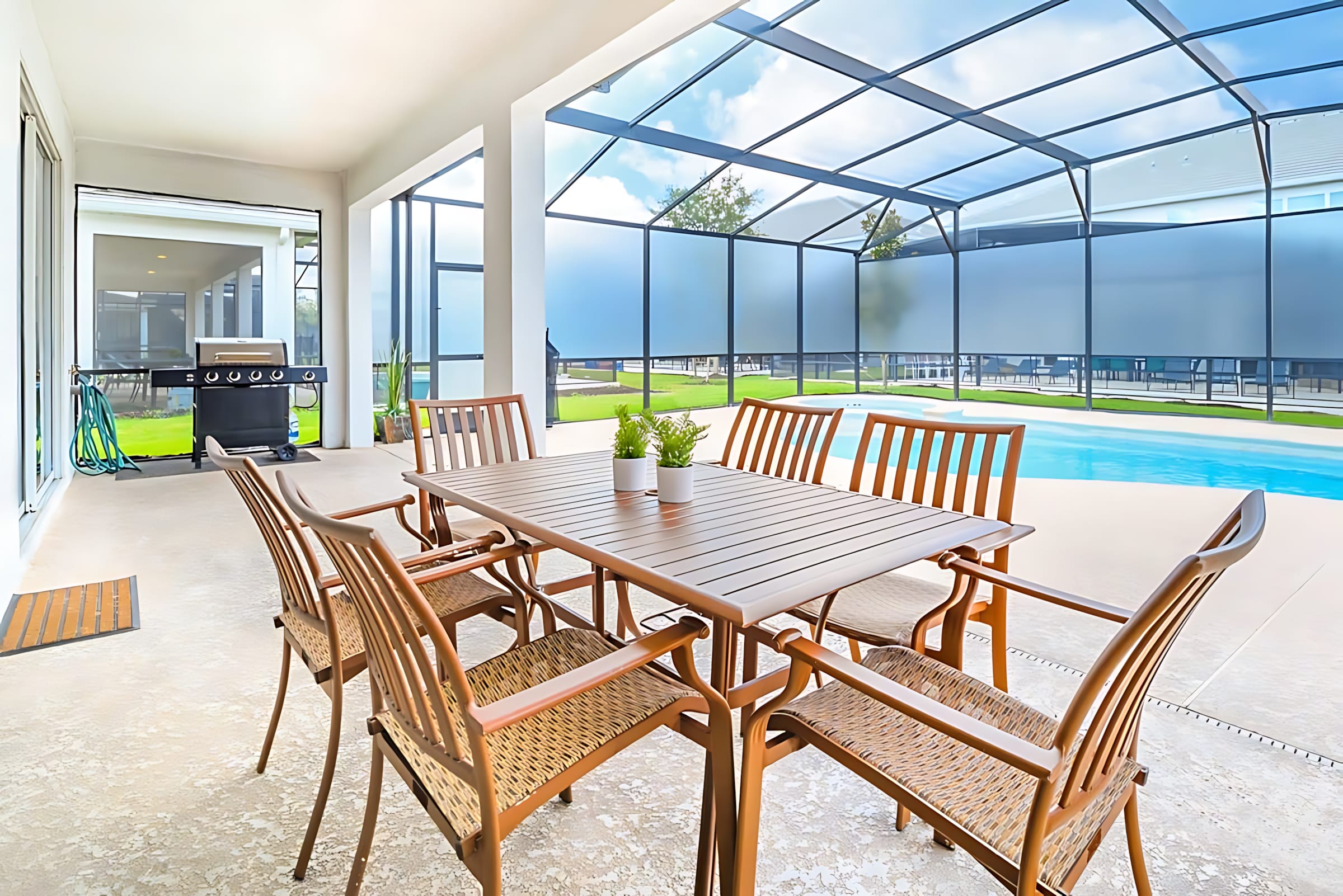 Covered outdoor patio with a wooden dining table and six chairs, a BBQ grill in the background, all within a screened enclosure overlooking a swimming pool.