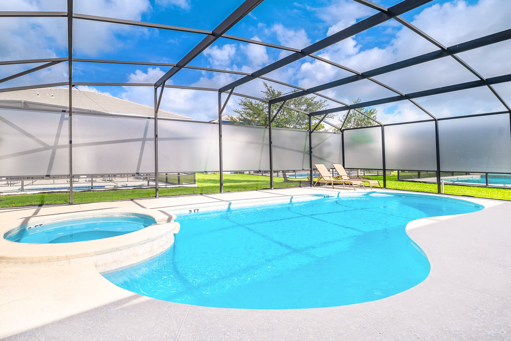Expansive screened pool area featuring a large freeform swimming pool and a circular hot tub, surrounded by a light patio deck with lounge chairs and a grassy area beyond the screen.