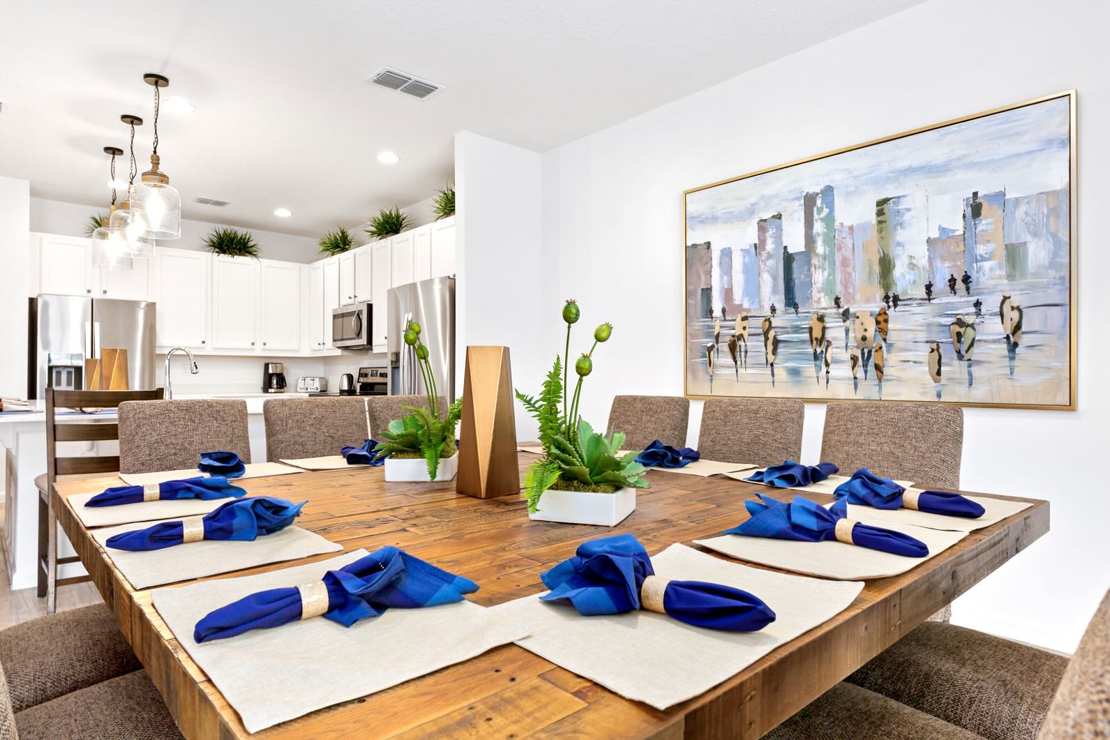 Dining room with a large square rustic wooden table and upholstered chairs, featuring a modern cityscape painting, and an open view to a white kitchen with stainless steel appliances in the background.