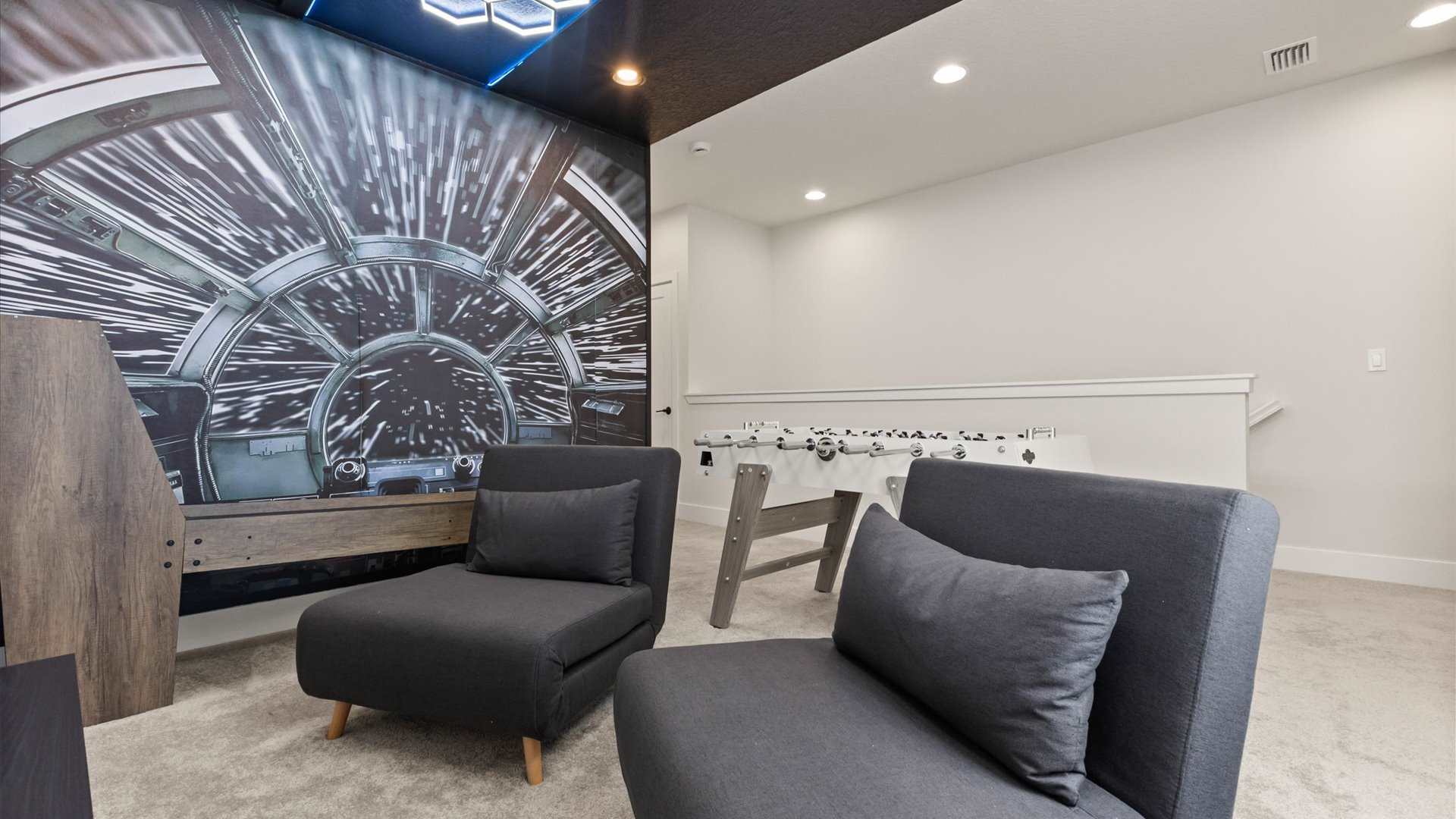 Cozy corner of the game room featuring two dark gray armchairs, a foosball table, and a striking spaceship cockpit wall mural. The room is carpeted and offers a comfortable spot for relaxation and play.