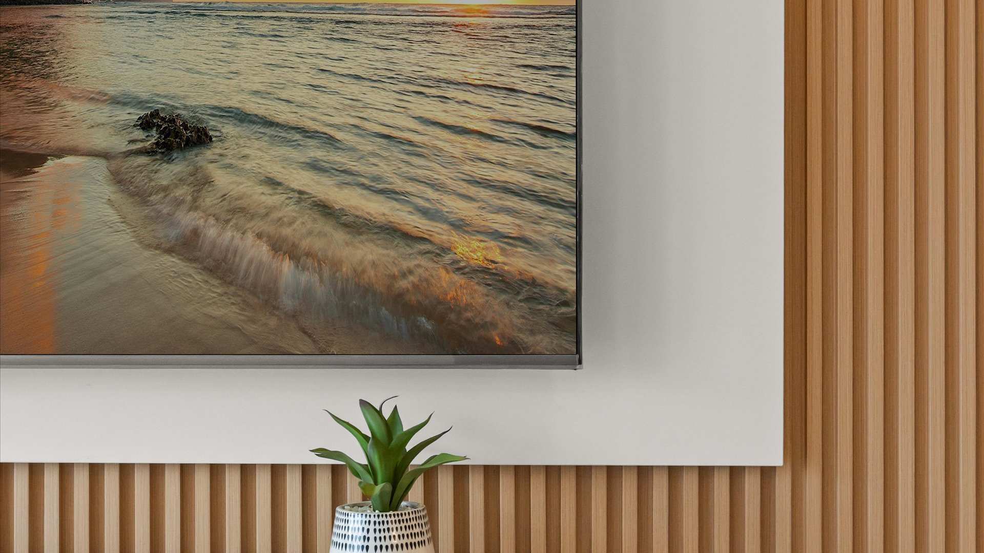 A close-up of the wall-mounted TV displaying a beach scene, set against a white panel and light wood slat accent wall, with a small green potted plant on the white floating console below.