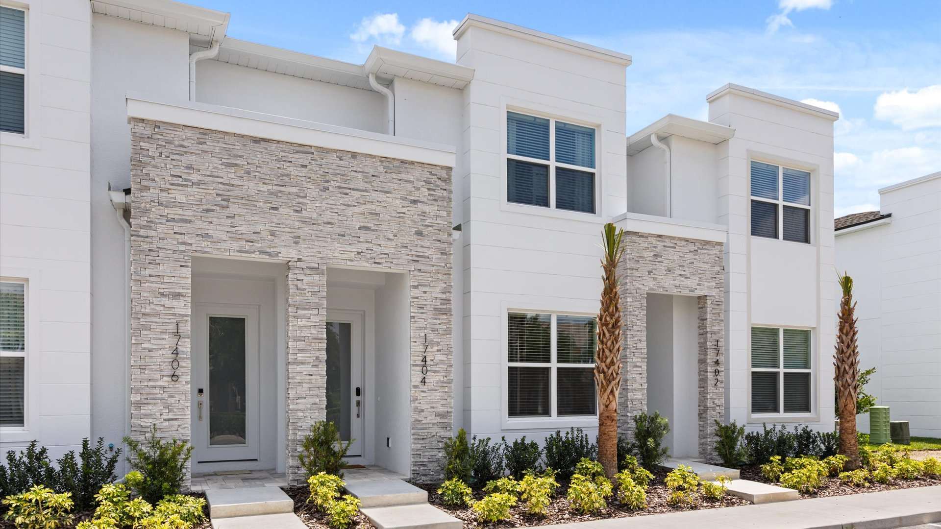 Modern townhouse exterior featuring white stucco and light gray stacked stone accents around the entrances. Large windows with blinds are visible, and small palm trees and shrubs line the front walkway.