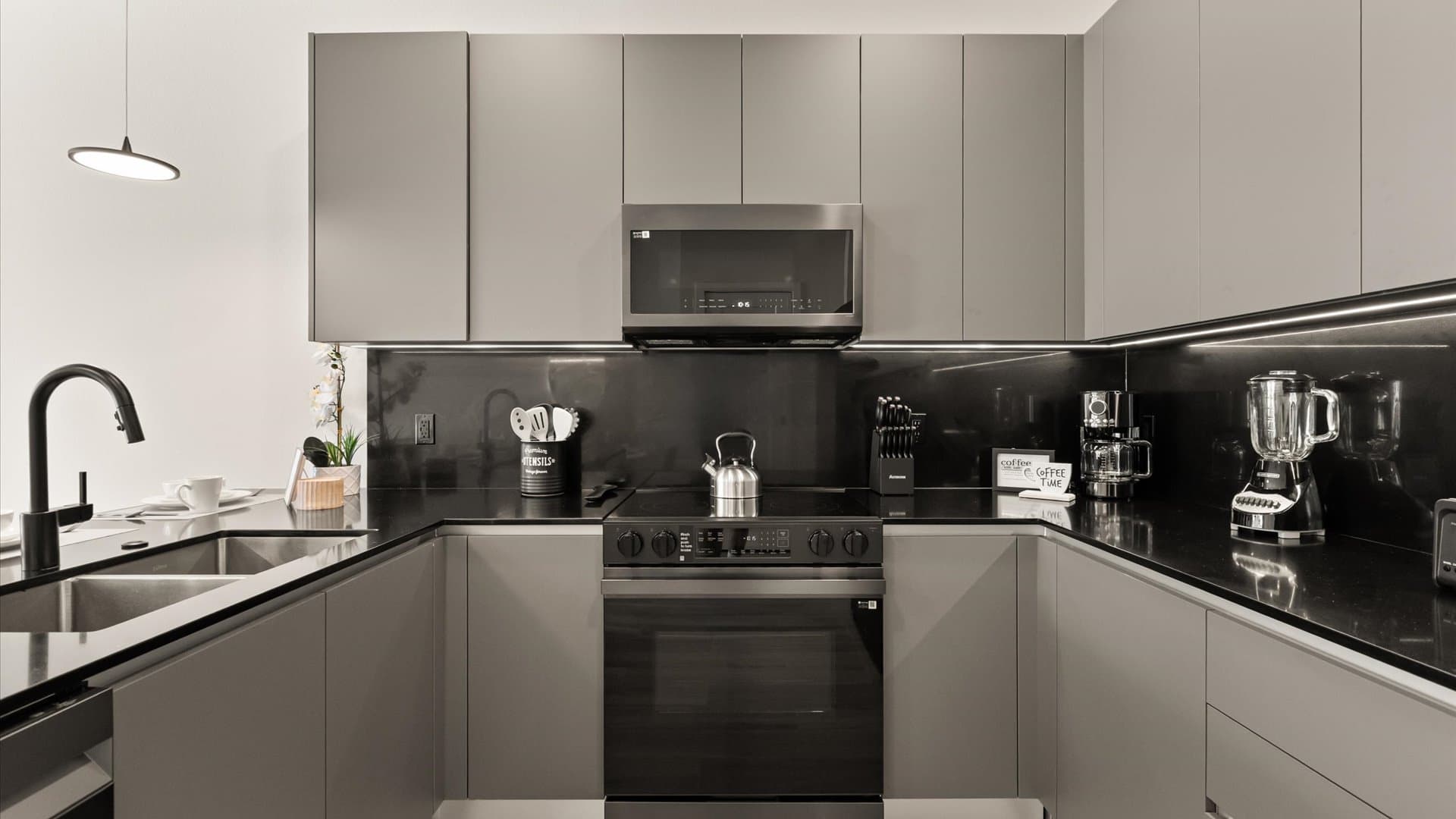 Sleek modern kitchen with gray flat-panel cabinets, black countertops and backsplash, and stainless steel appliances including a microwave, stove, and sink, illuminated by under-cabinet lighting.