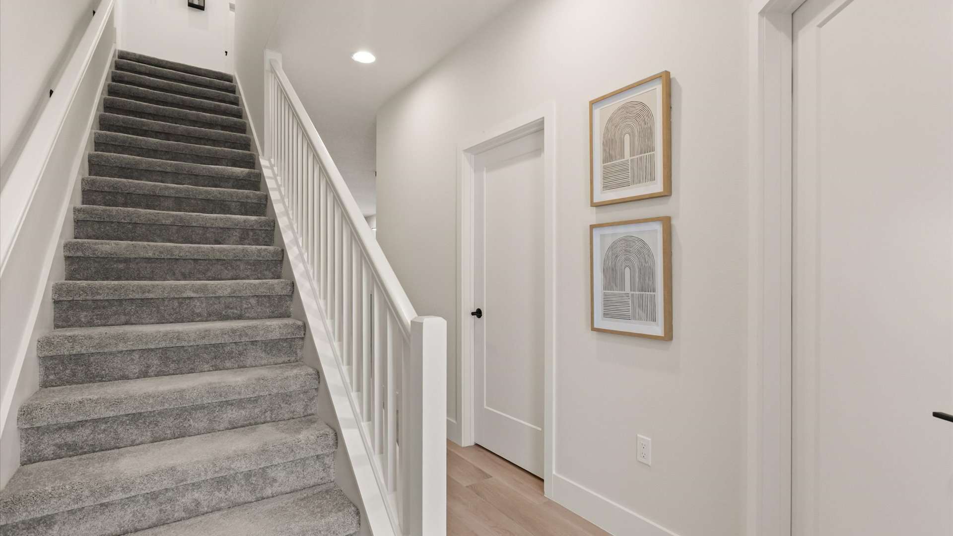 A modern white hallway features a carpeted staircase with white railings on the left, and two framed abstract art pieces hanging on the right wall above light wood flooring.