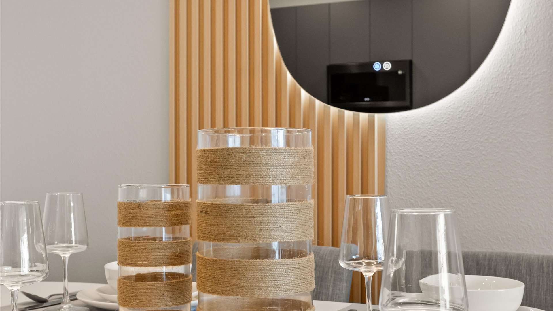 A close-up of the dining table centerpiece, featuring two glass vases wrapped with natural twine, surrounded by wine glasses and white dinnerware, with a modern illuminated mirror and wood slat wall in the background.
