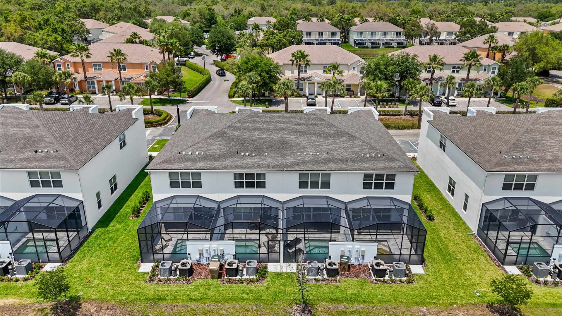 An aerial view showcasing the rear of several white townhouses, each featuring a private screened swimming pool in its backyard. The properties are separated by green lawns, with air conditioning units lined up behind each house, and a residential street visible in the distance.