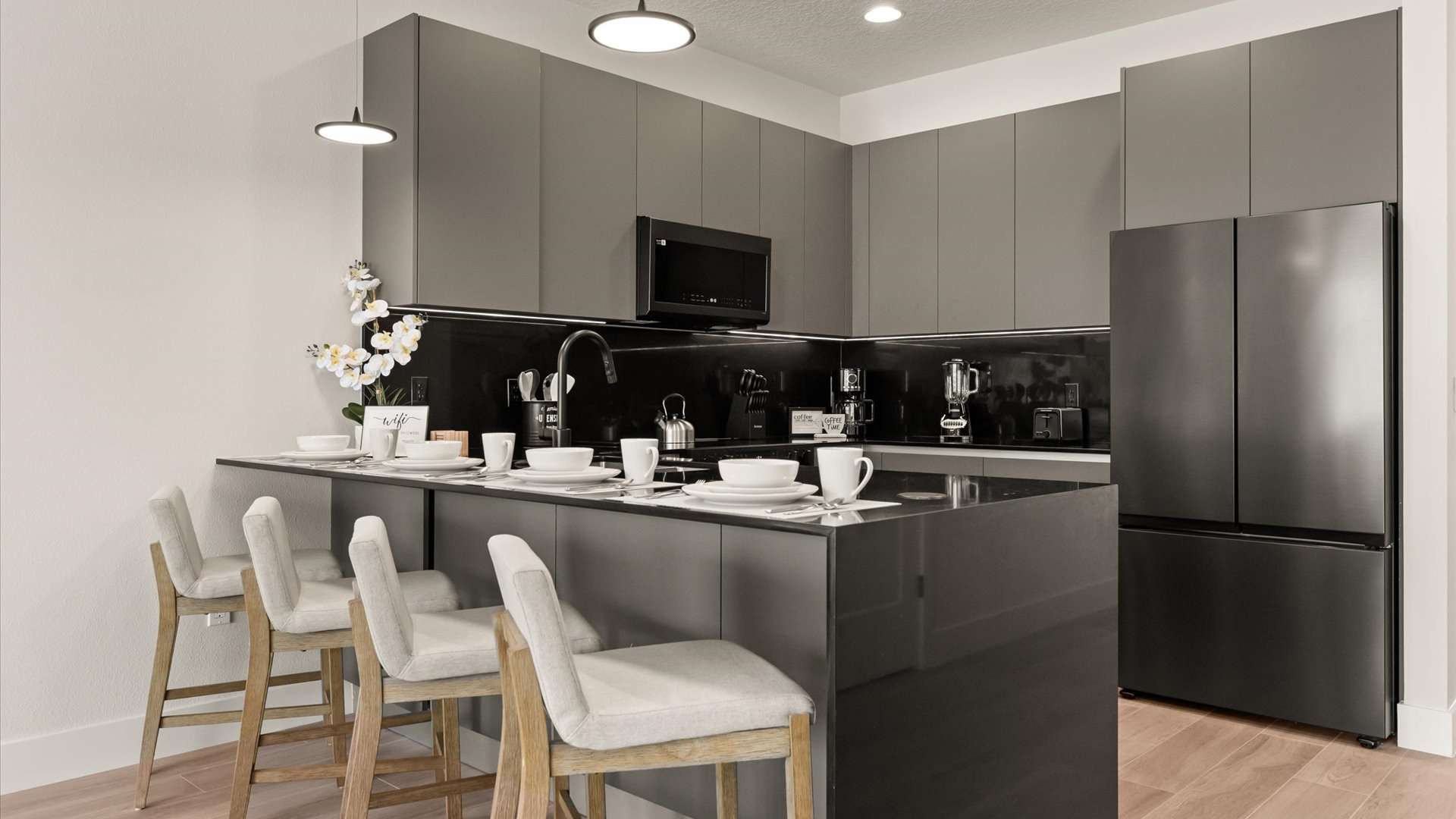 A close-up of the modern dark gray kitchen, highlighting the island with a sink and breakfast bar set with white dishes, alongside sleek cabinetry, a black microwave, and a large black refrigerator.
