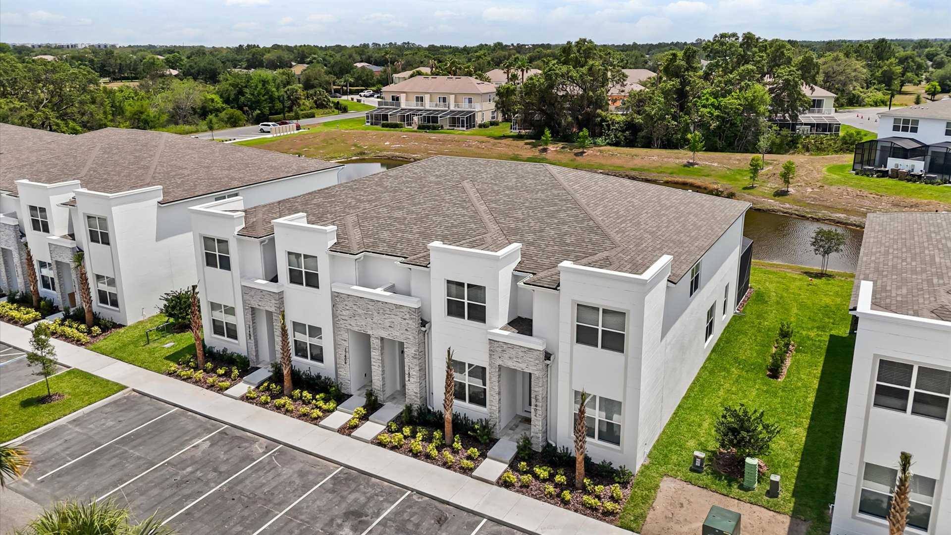 An aerial view shows a row of modern white townhouses with gray roofs, surrounded by green lawns, a parking lot, and a pond in the background.