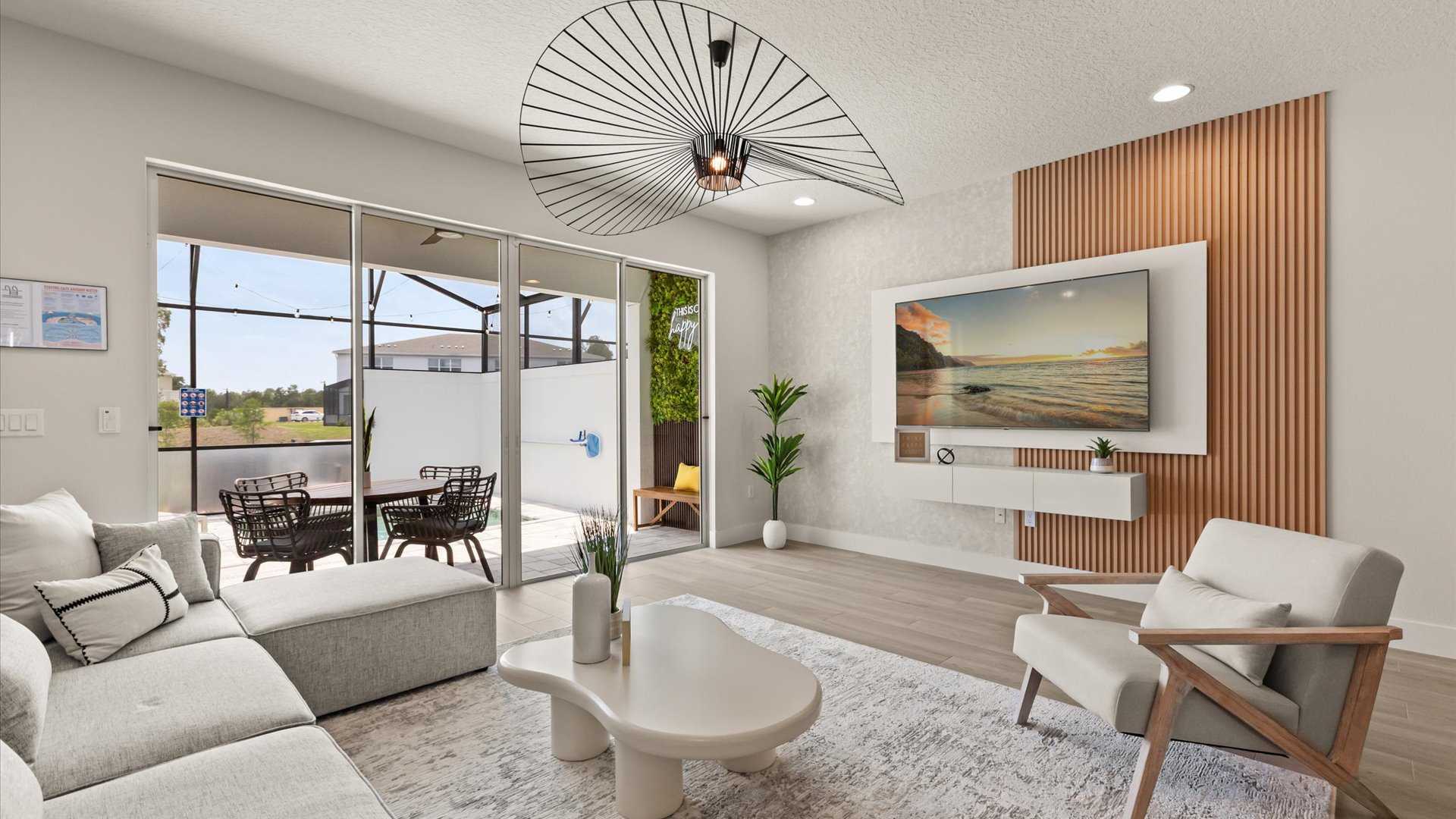 Bright living room with a light gray sectional sofa, a white organic coffee table, and light wood flooring. A large sliding glass door opens to a screened patio, and a wall-mounted TV is set against a slatted wood accent wall.