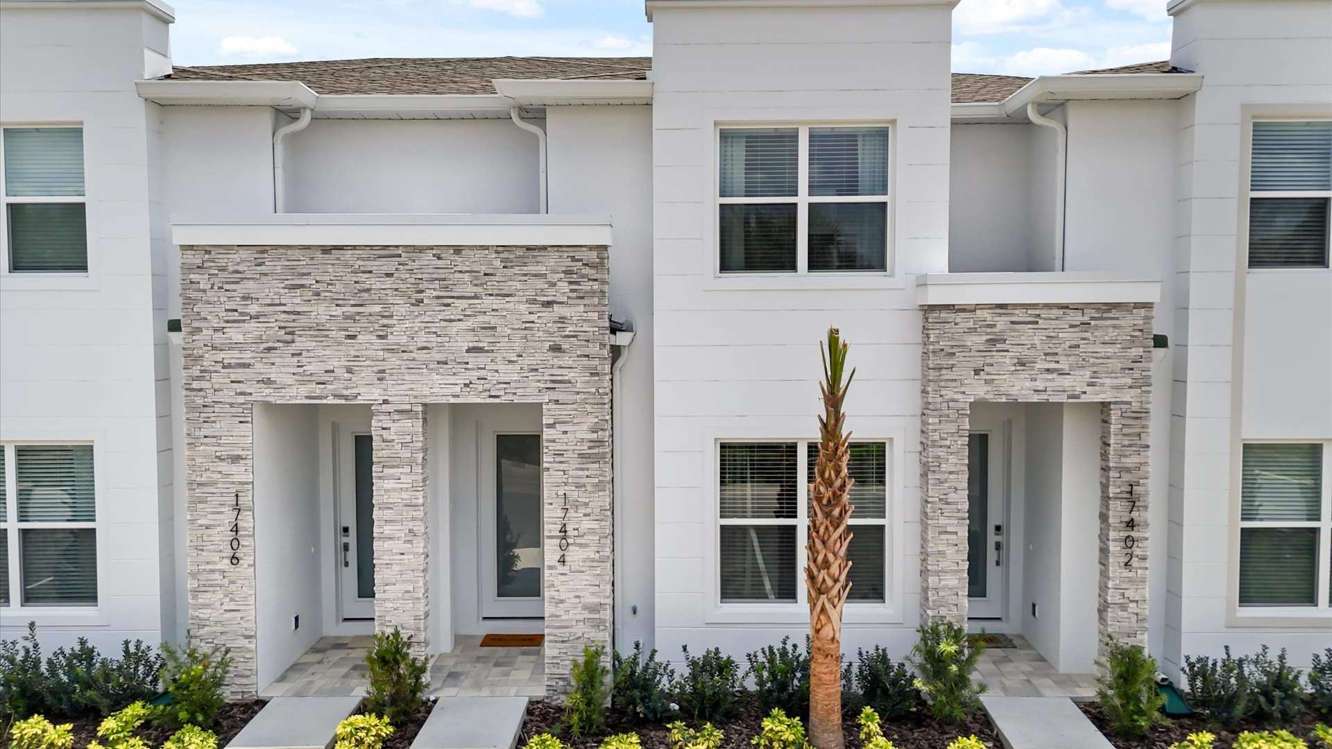 The front exterior of two modern white townhouses, each with a stone-clad entrance, large windows with blinds, and small palm trees and shrubs in front.
