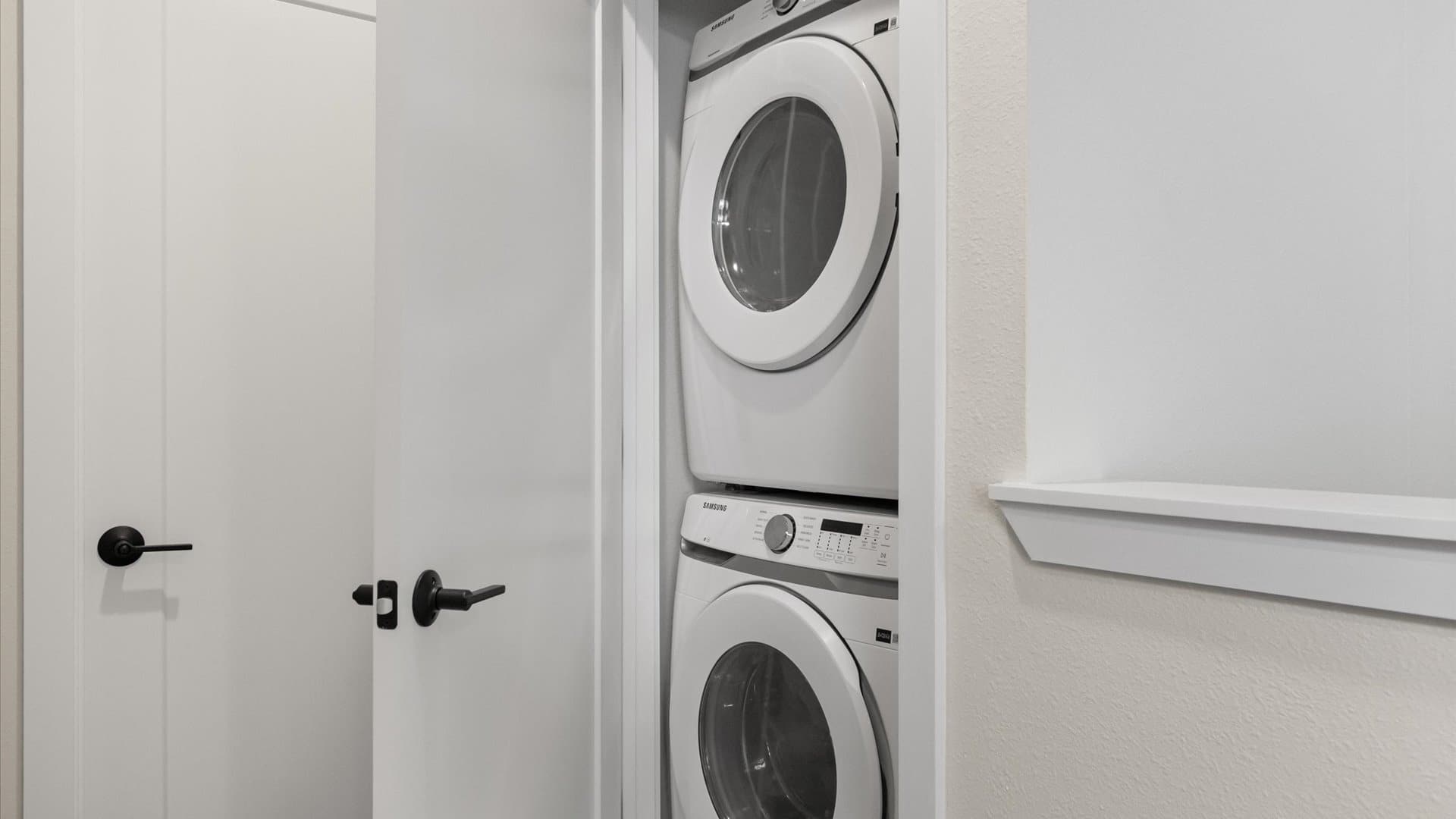 A compact laundry closet features a stacked white Samsung washer and dryer unit, with white doors and black handles on the left.