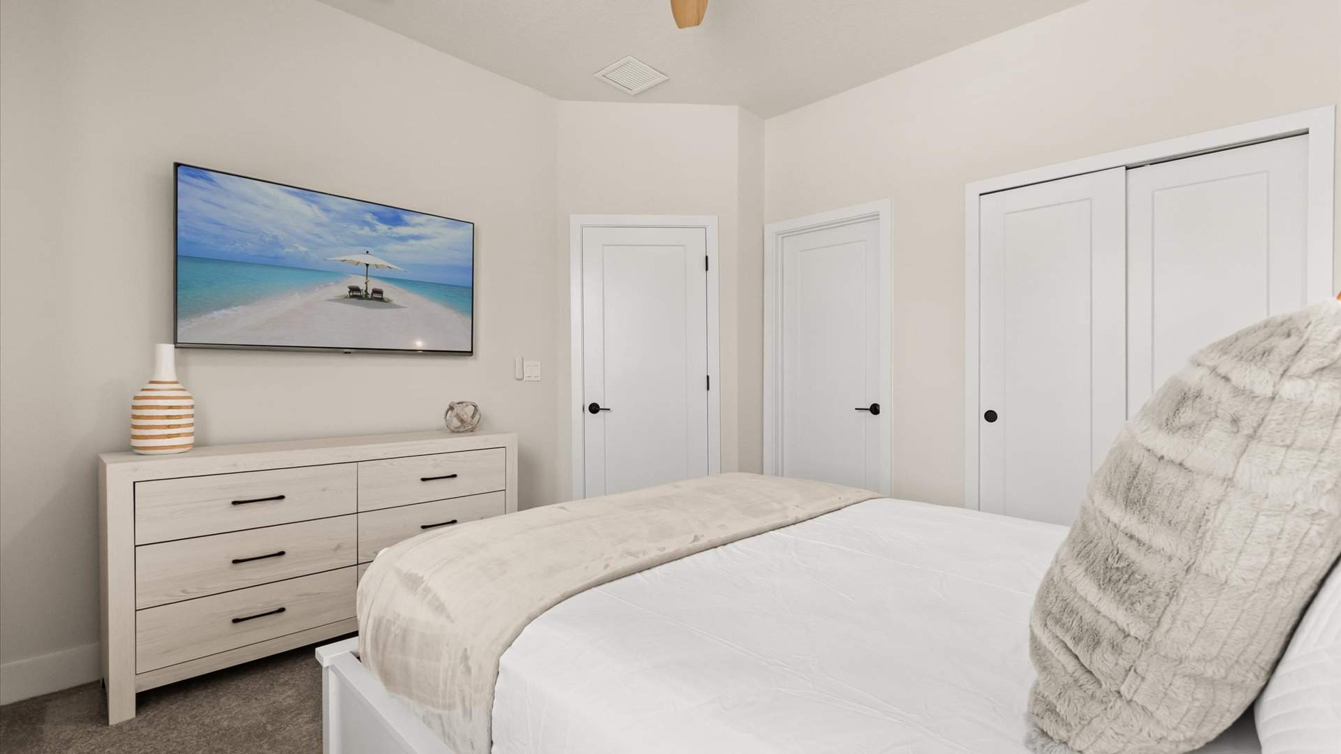 Serene bedroom featuring a white wooden bed with light bedding, a matching dresser with black handles, and a wall-mounted TV displaying a beach scene, all on a light carpeted floor.